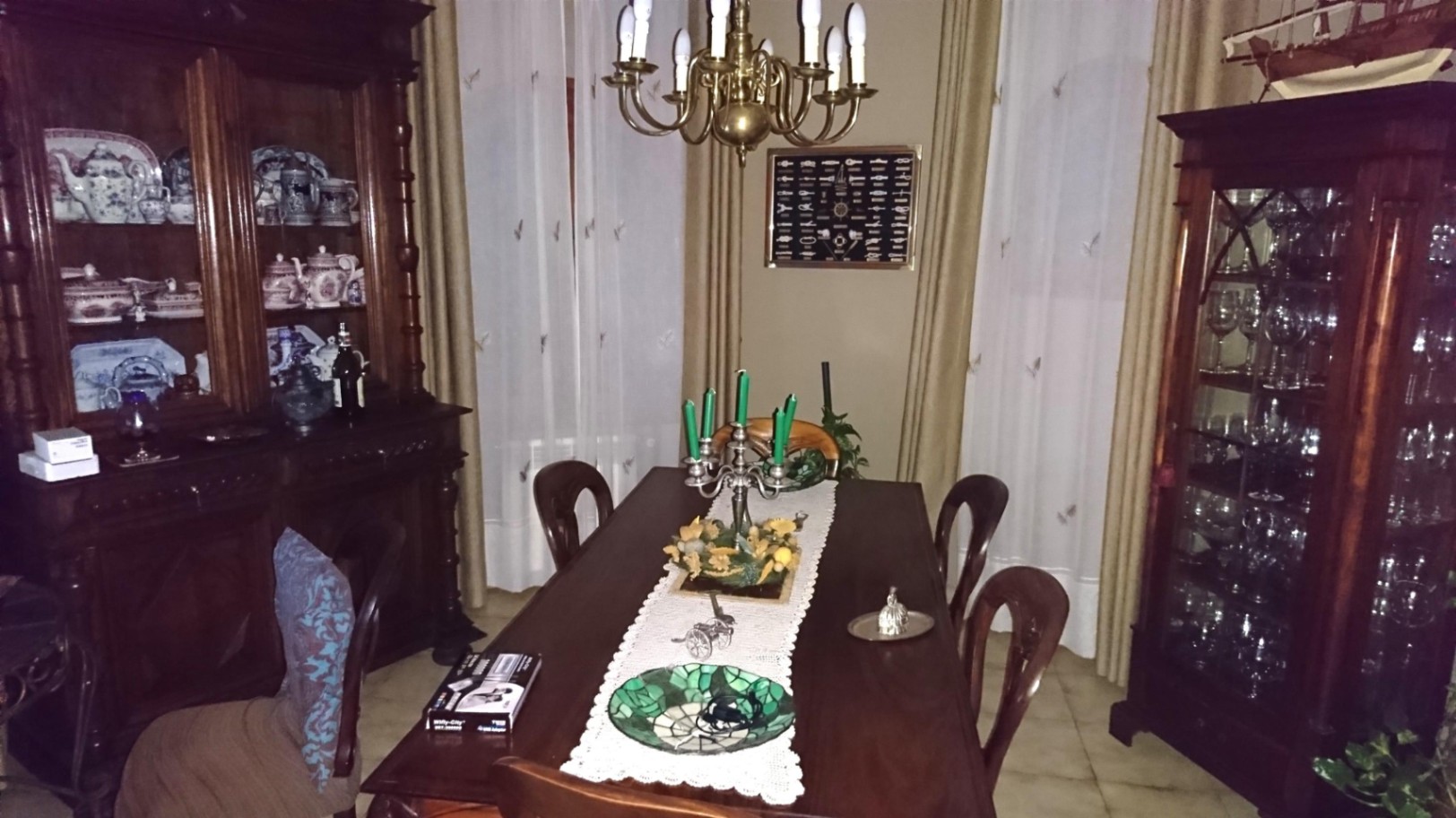 Comedor de estilo clásico con mesa de madera oscura, vitrinas ornamentadas para vajilla y una lámpara de araña de latón sobre textiles elegantes.