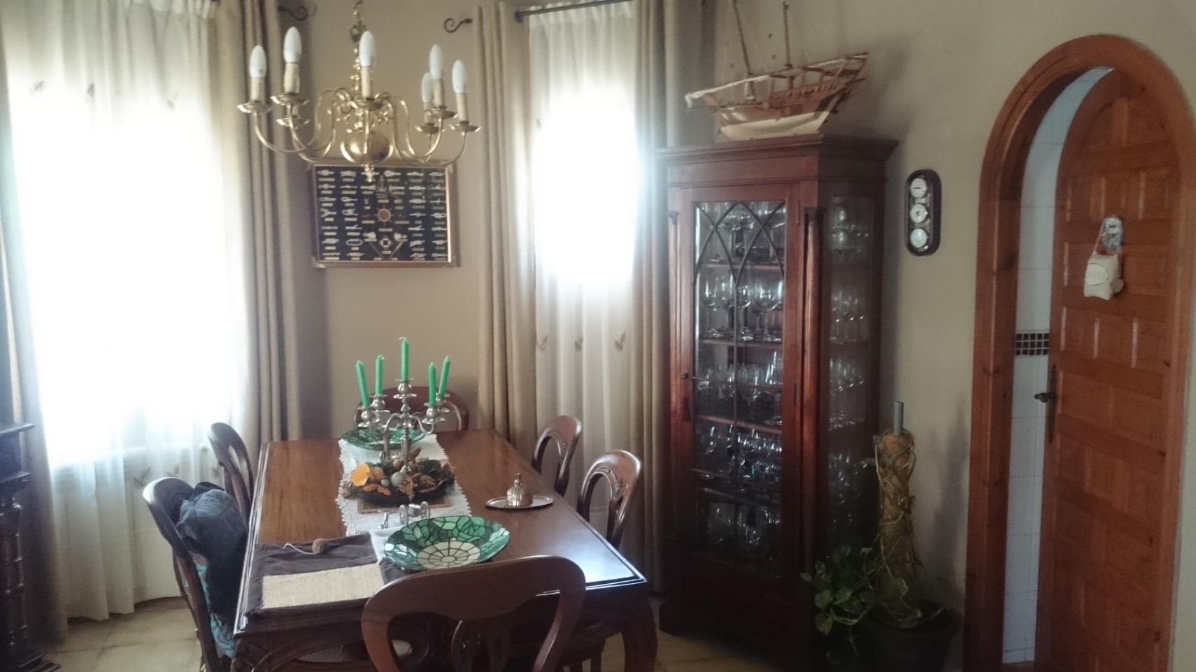 Comedor tradicional con mobiliario de madera, vitrina para cristalería y lámpara de latón. Incluye un acceso mediante arco de madera y luz natural filtrada.