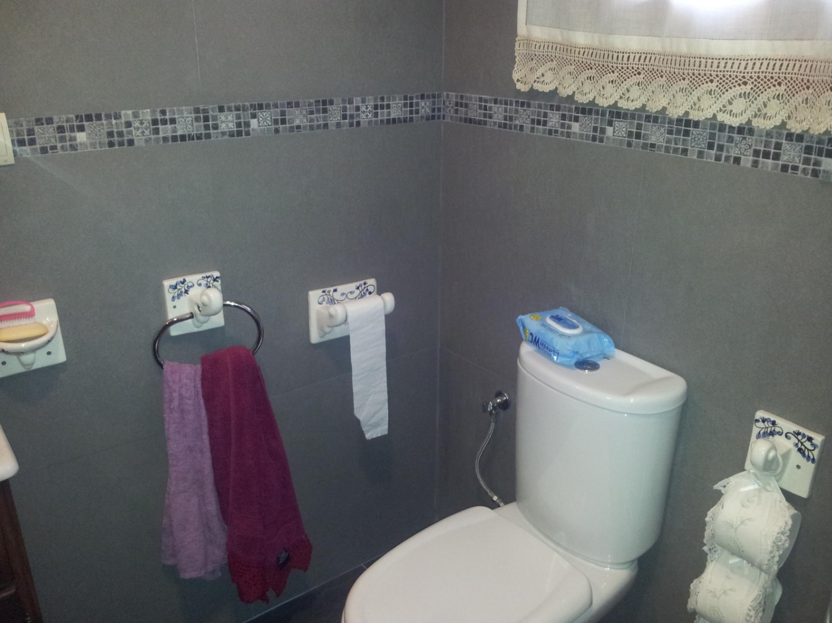 Cuarto de baño con azulejos grises y cenefa de mosaico, equipado con sanitarios blancos y accesorios cerámicos decorados con motivos florales azules.