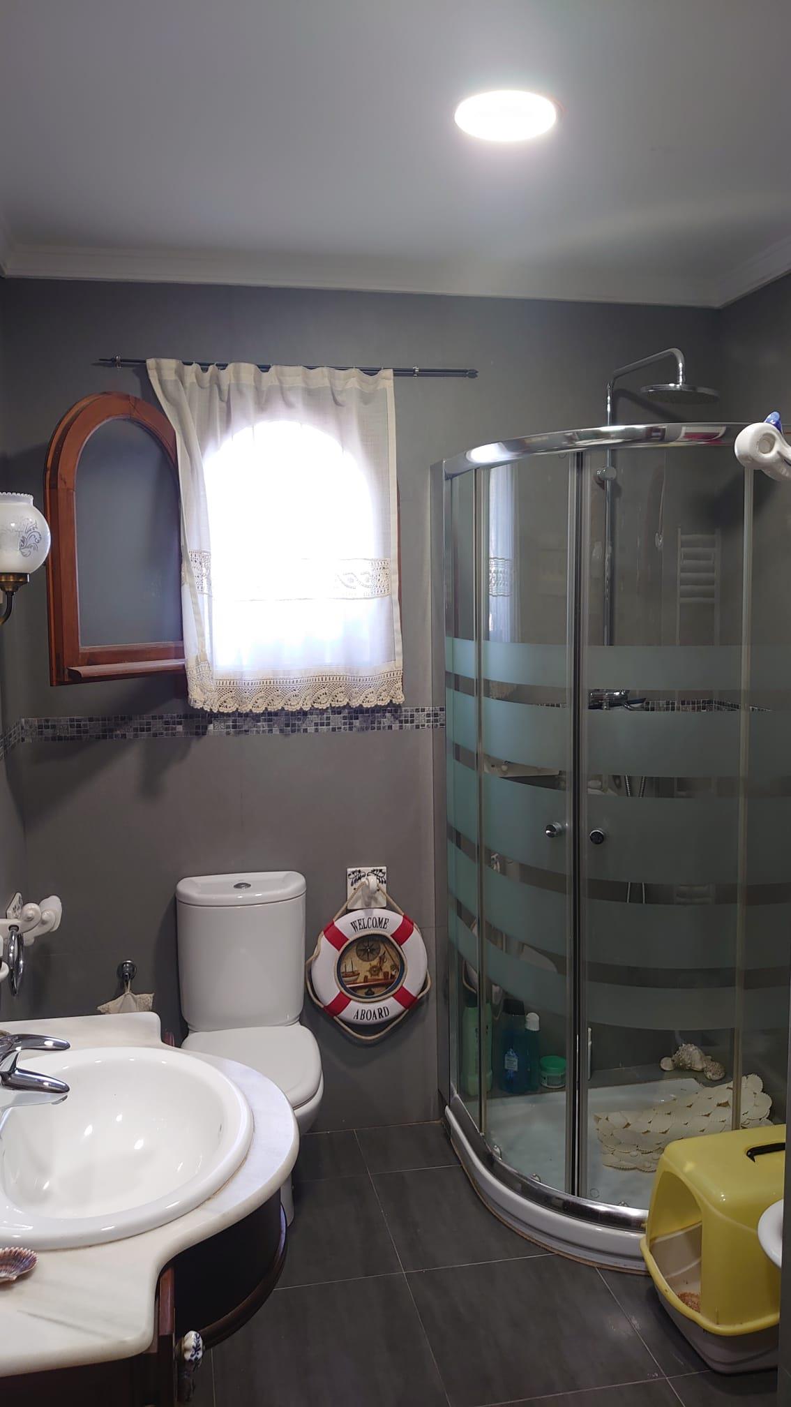 Cuarto de baño con ducha de esquina acristalada, paredes en tono gris y encimera de mármol. Incluye una ventana de arco con madera y decoración de estilo náutico.