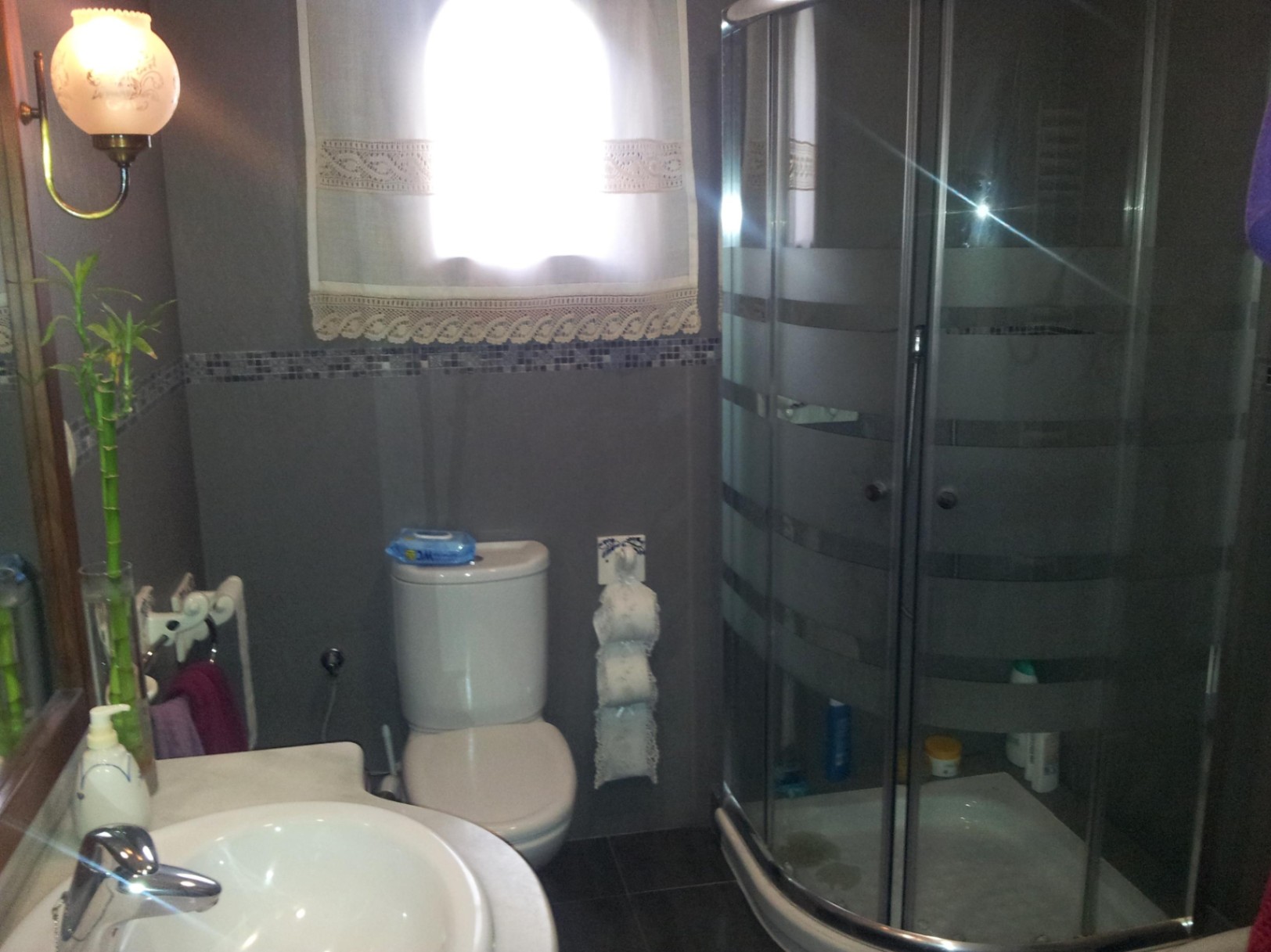 Cuarto de baño con ducha de esquina de cristal curvado, paredes en tonos grises con cenefa de mosaico y un lavabo blanco integrado.
