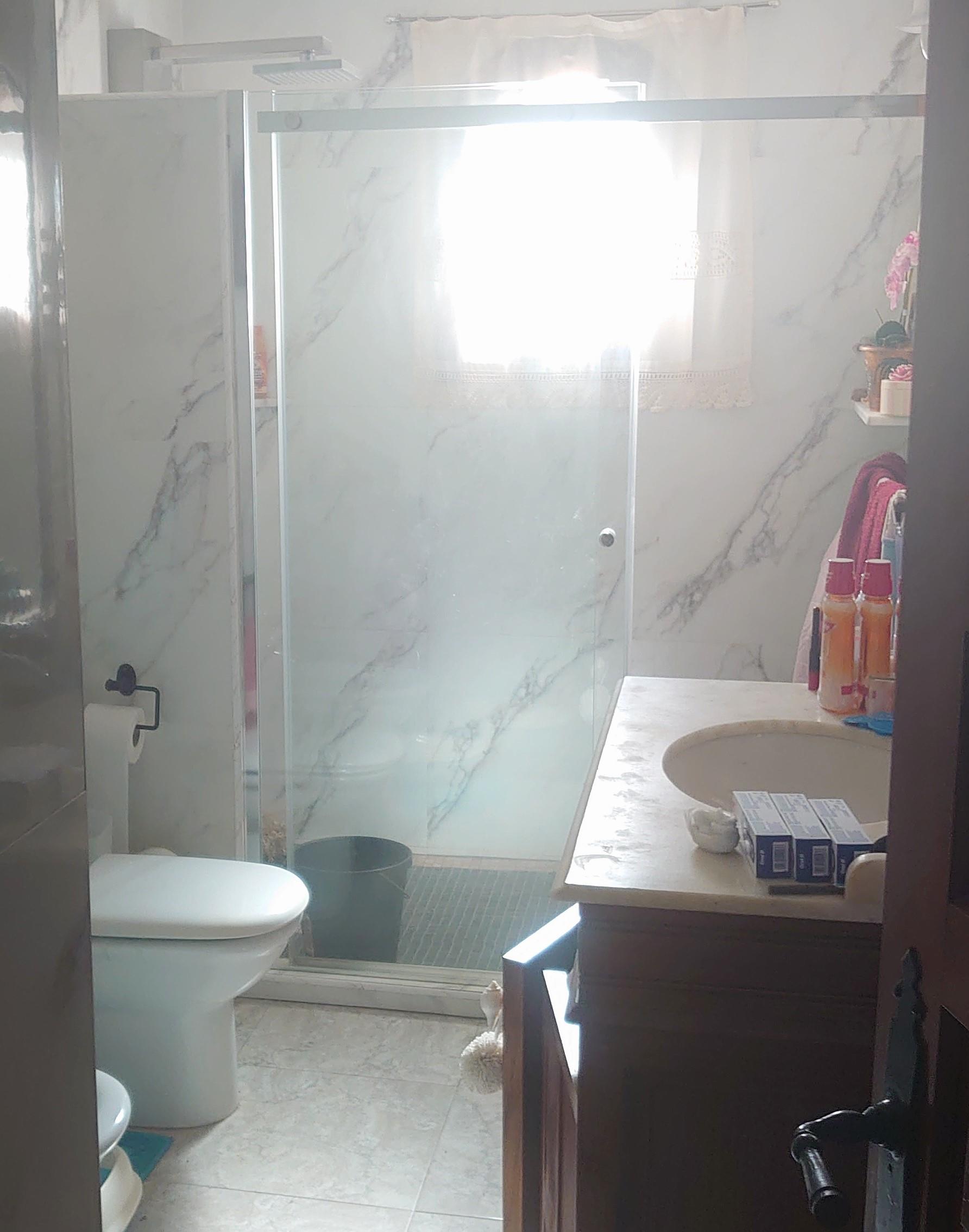 Cuarto de baño con ducha de obra, mampara de cristal y paredes revestidas en mármol veteado. Incluye un mueble de lavabo en madera con encimera de piedra natural.
