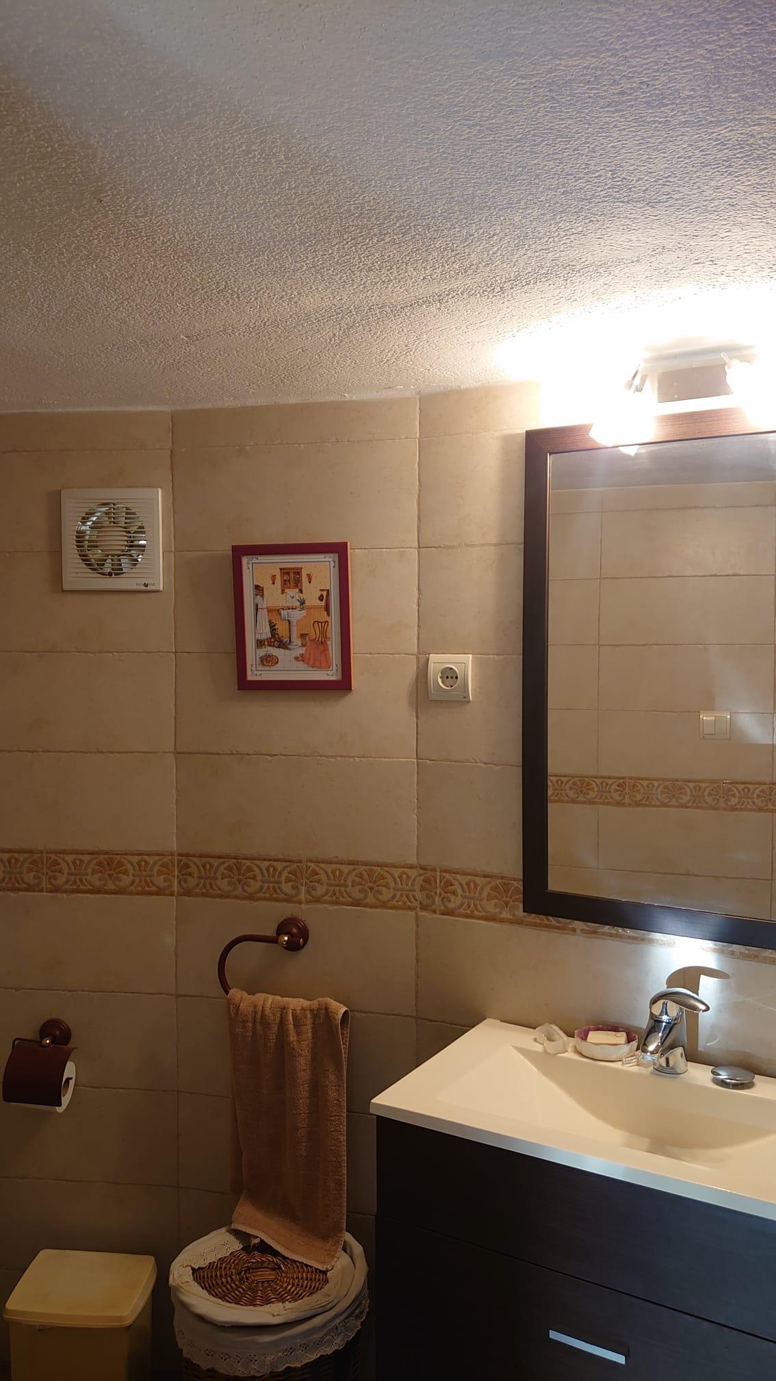 Cuarto de baño con lavabo integrado sobre mueble oscuro, espejo enmarcado con iluminación superior y paredes alicatadas en tonos beige con cenefa decorativa.