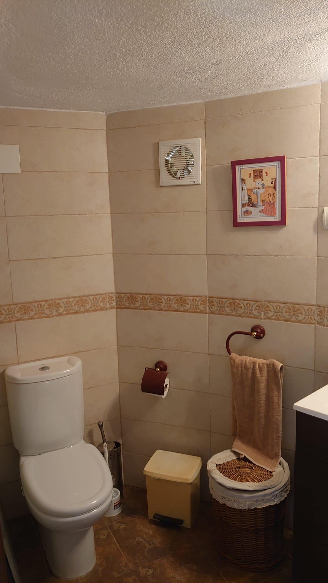 Cuarto de baño funcional con azulejos en tonos crema, cenefa ornamental y sanitarios blancos. Dispone de accesorios en madera oscura y un cesto de mimbre.