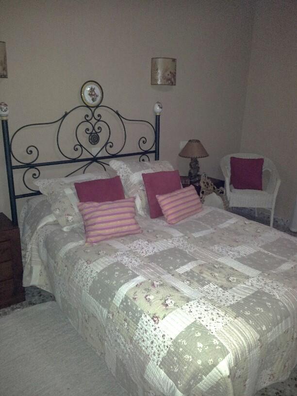 Dormitorio con cabecero de hierro forjado, colcha de estilo patchwork y cojines en tonos rosa. Incluye un sillón de mimbre blanco y apliques de pared decorativos.