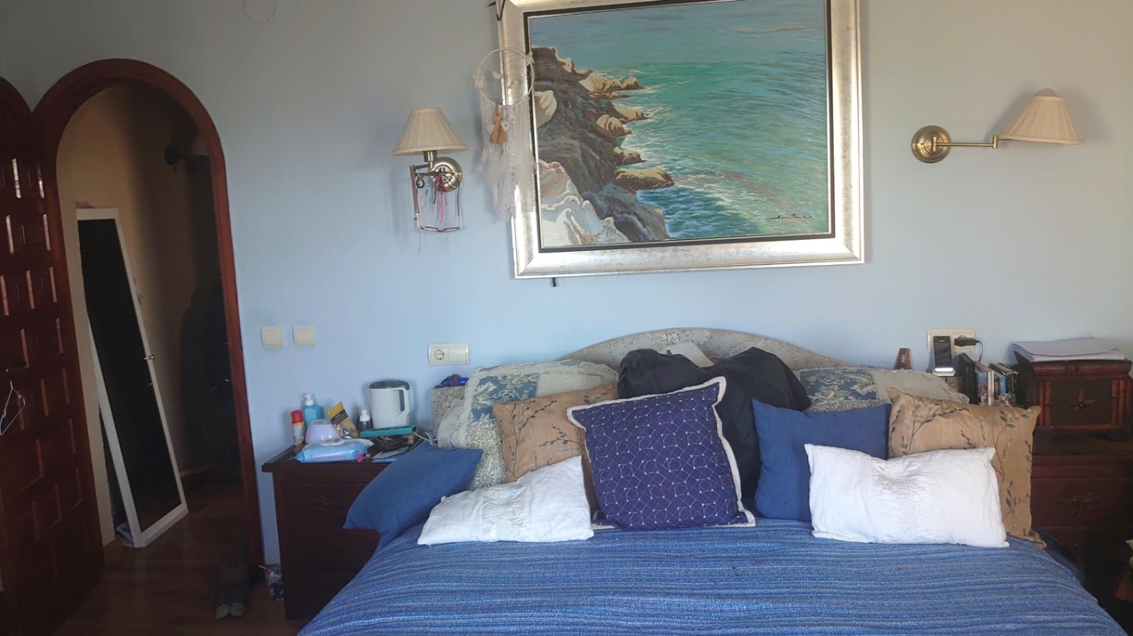 Dormitorio con paredes azul suave, cama con textiles variados, cuadro costero central y acceso mediante un arco de madera tradicional.