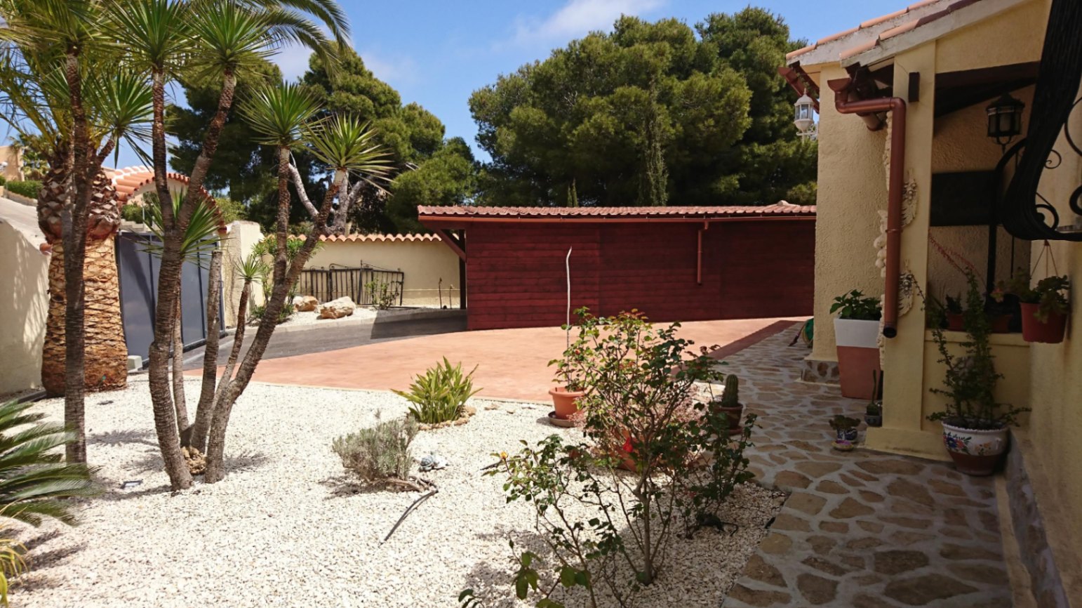 Entrada exterior con camino de piedra natural, zona de aparcamiento pavimentada y jardín de bajo mantenimiento con palmeras y grava blanca.
