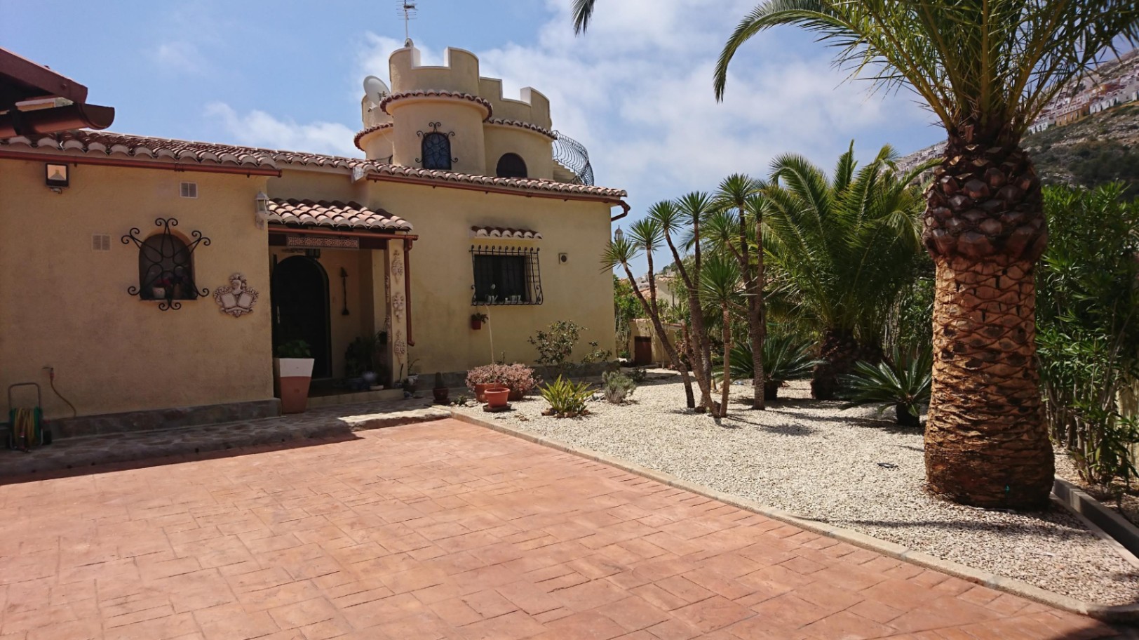 Fachada mediterránea con torreón almenado, patio de baldosas de terracota y jardín de grava con palmeras y yucas bajo el sol de Cumbre del Sol.