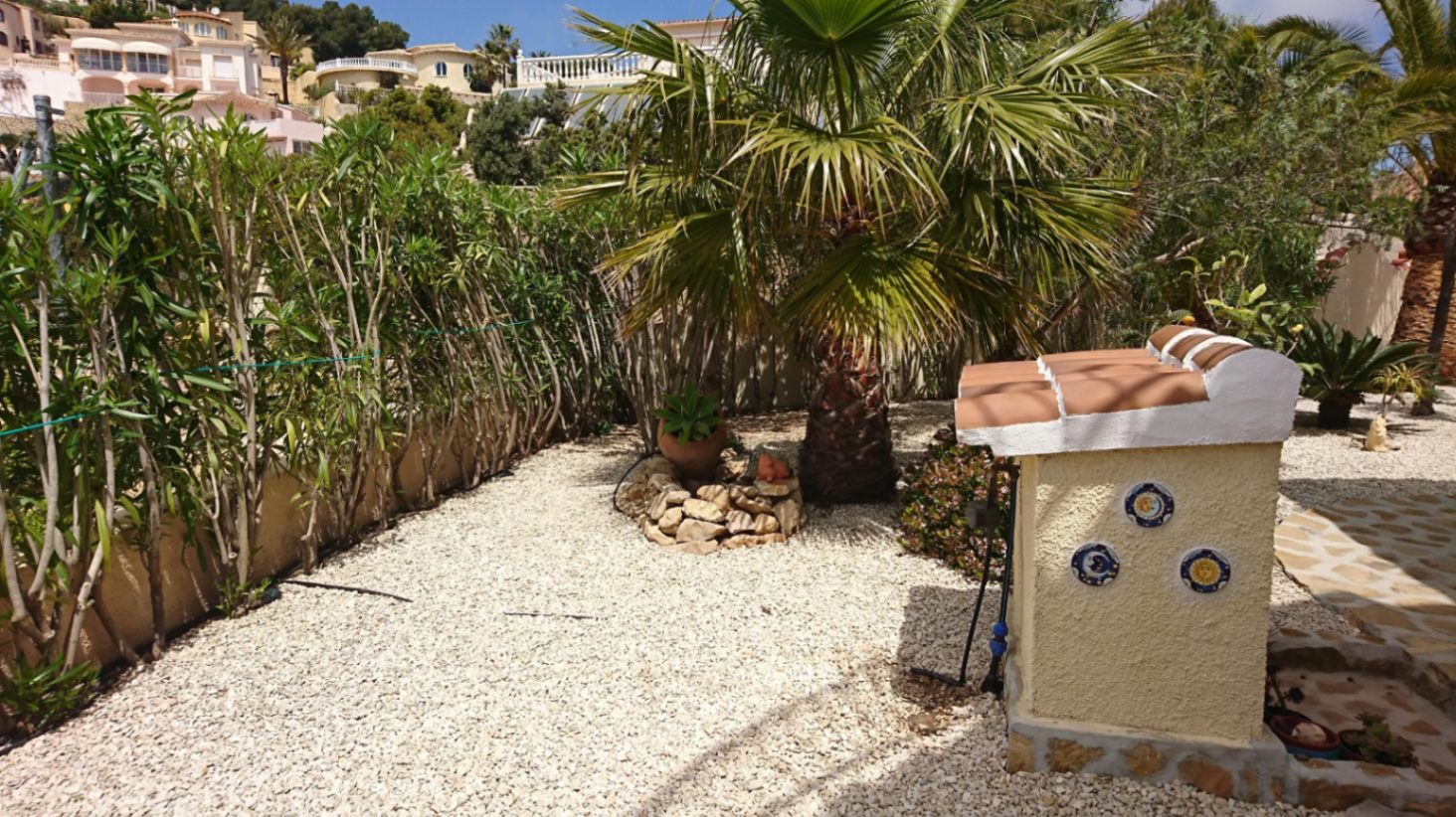 Jardín mediterráneo con suelo de grava blanca, una palmera central y un pequeño murete decorativo con tejas y platos cerámicos tradicionales.