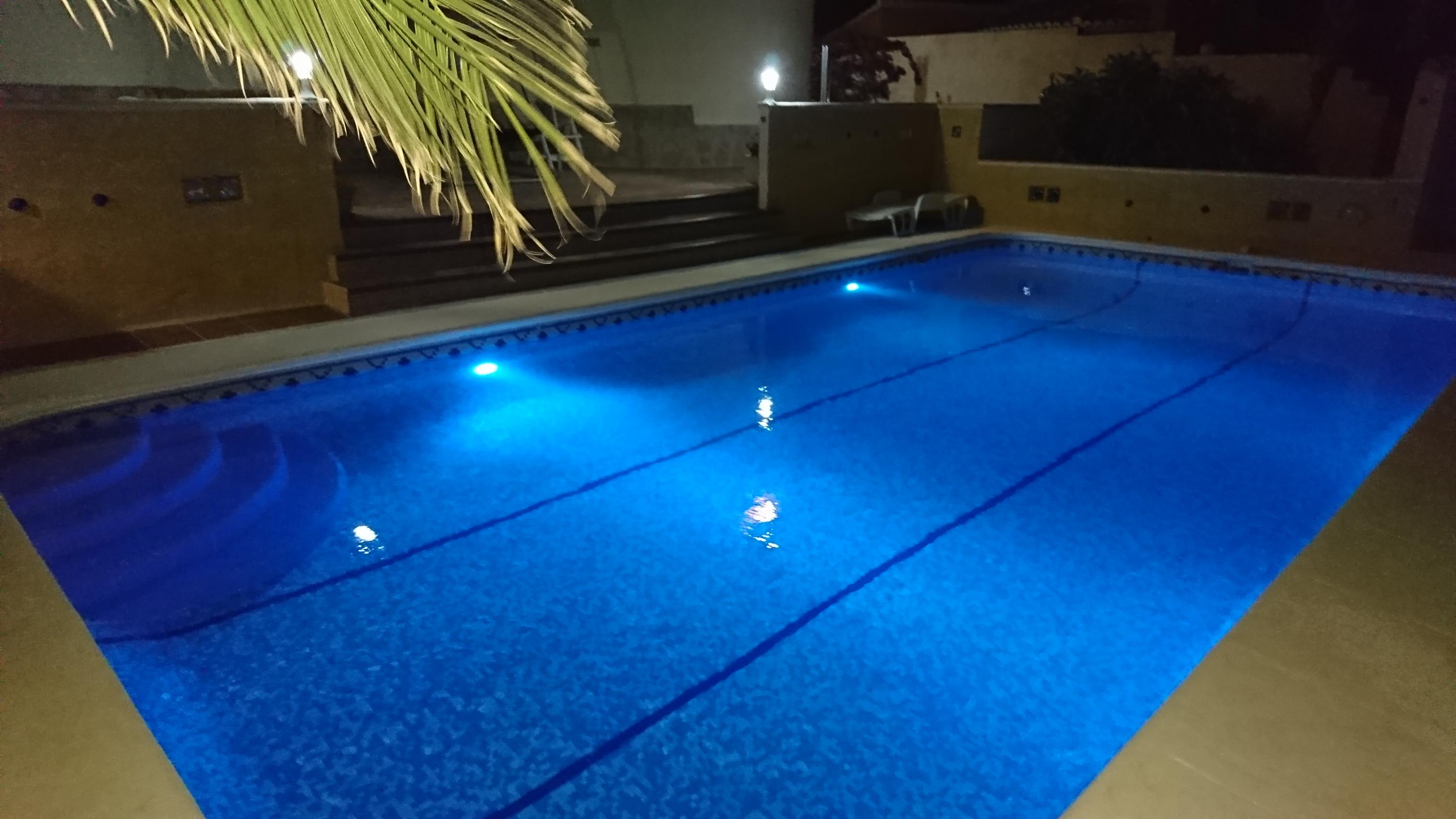 Piscina rectangular de 10x4 metros con iluminación LED azul, escaleras romanas integradas y terraza perimetral de piedra bajo iluminación nocturna.