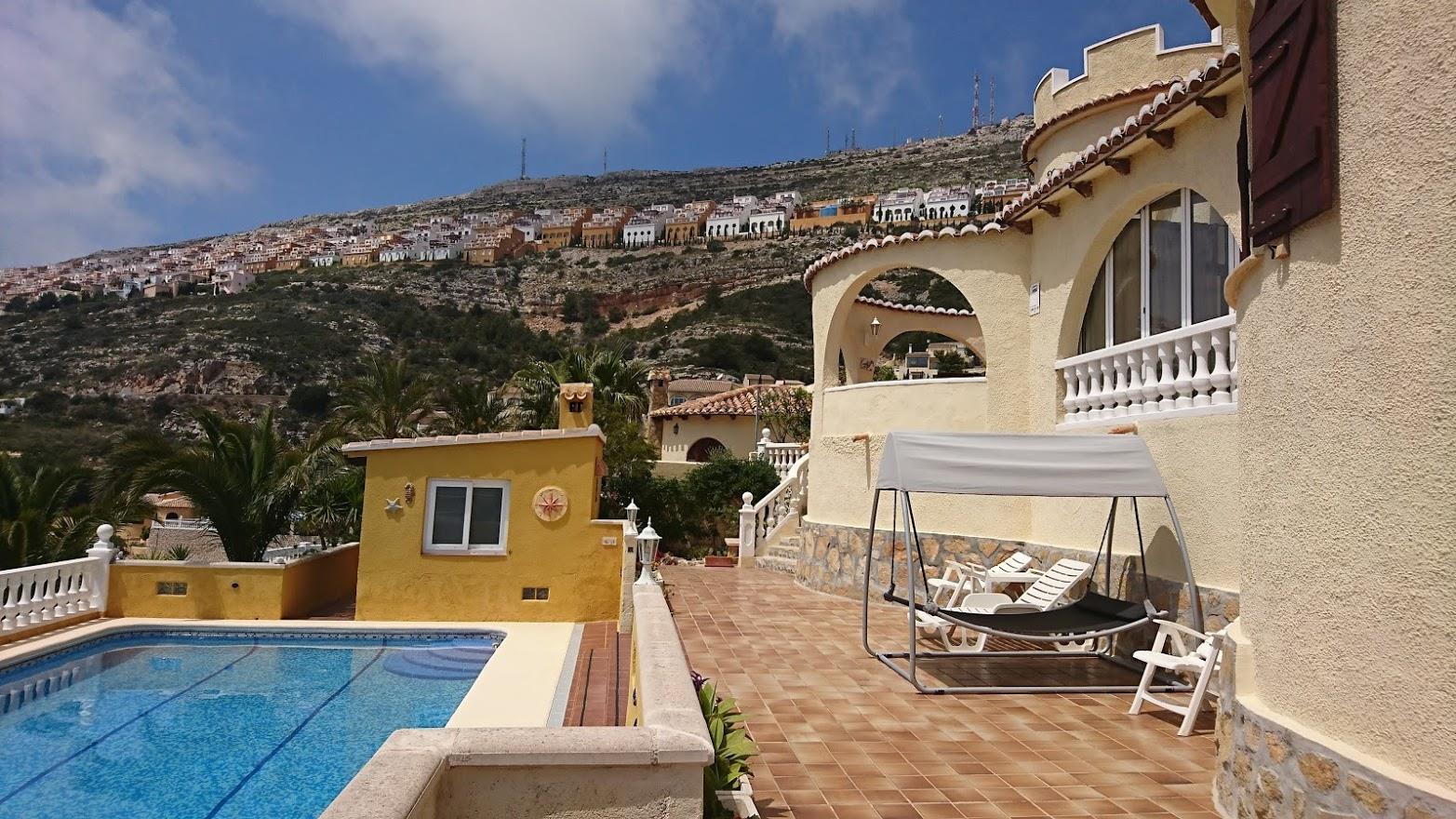 Terraza con piscina y mobiliario de exterior frente a la fachada de la villa, que muestra arcos tradicionales y una torre bajo la ladera de Cumbre del Sol.