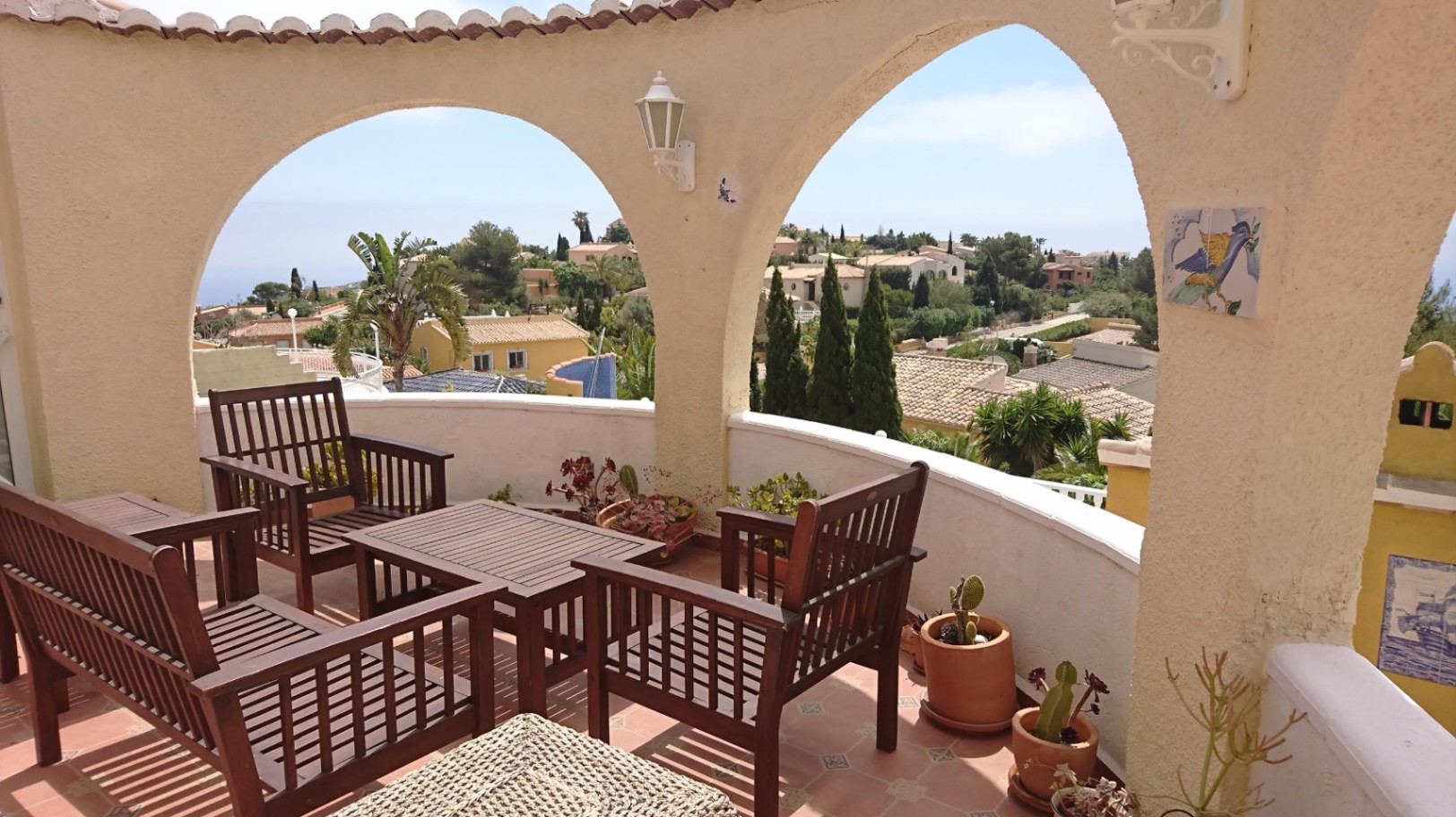 Terraza cubierta con arcos apuntados, mobiliario de madera oscura y suelos cerámicos. Ofrece vistas despejadas a la urbanización y al mar Mediterráneo.