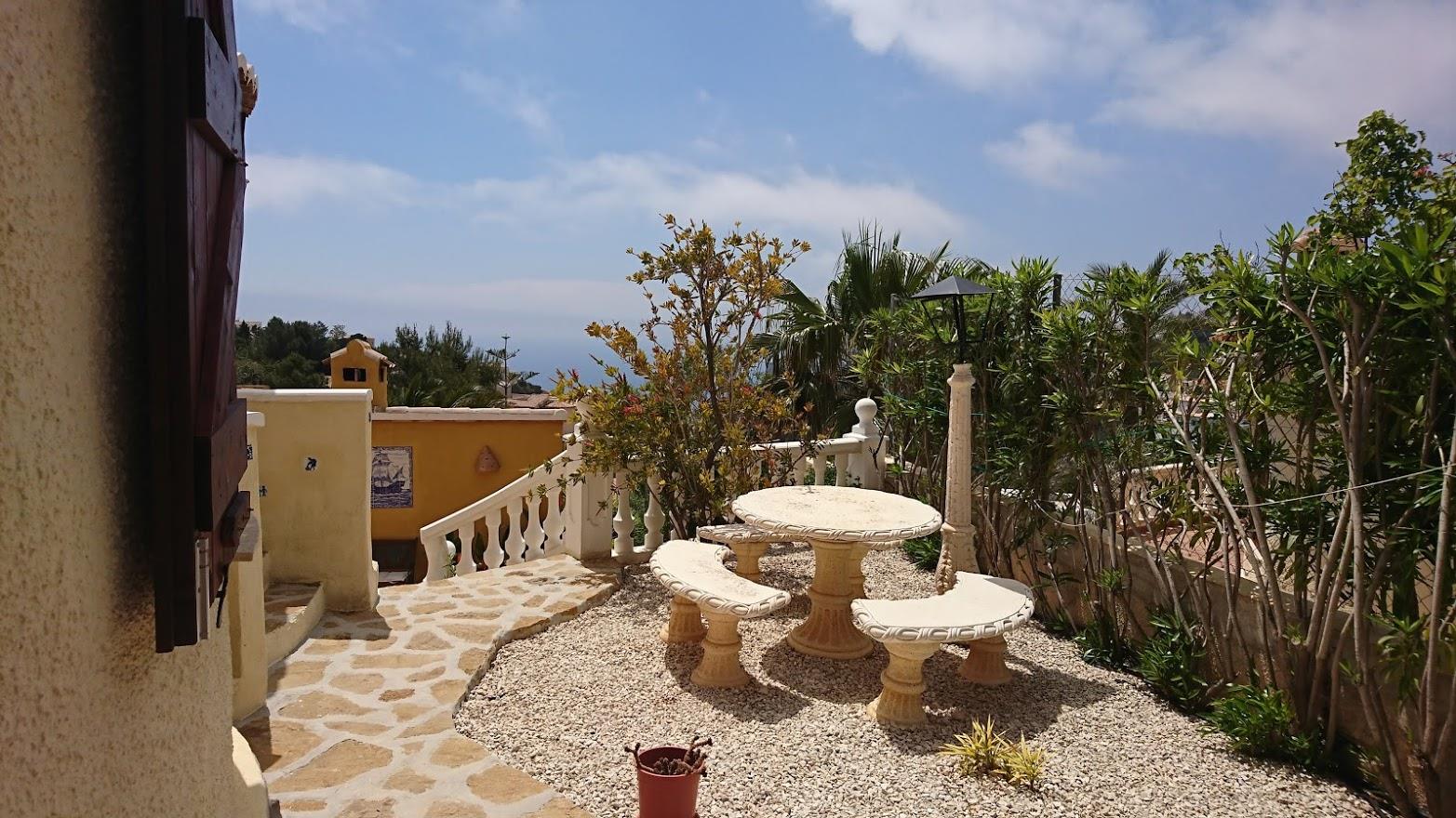 Terraza de grava con conjunto de mesa y bancos de piedra, delimitada por una balaustrada blanca y vegetación autóctona con vistas al Mediterráneo.