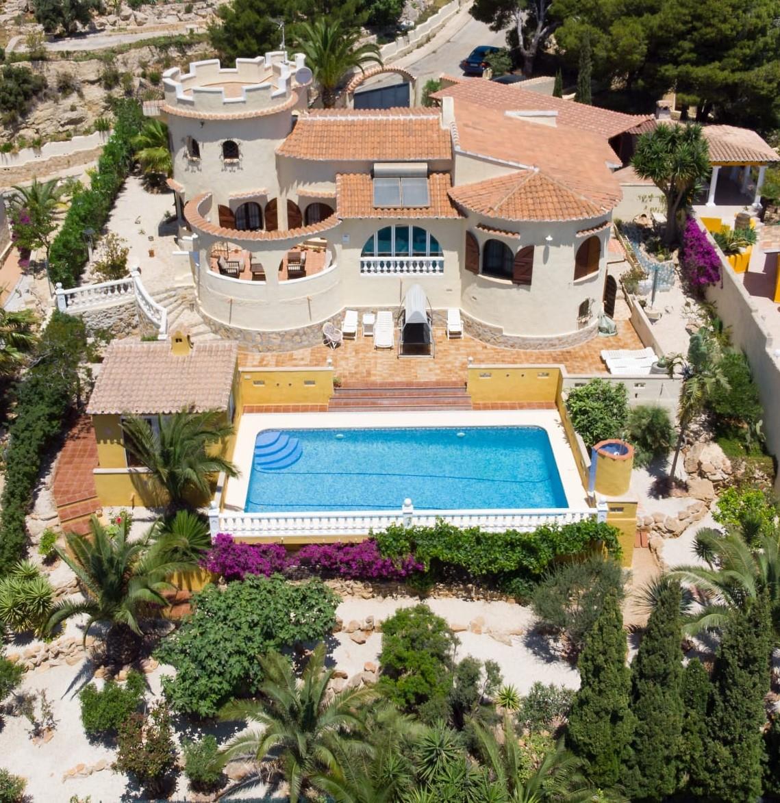 Vista aérea de la villa con arquitectura mediterránea, destacando su torreón almenado, piscina rectangular de 10x4m y jardines con palmeras y buganvillas.
