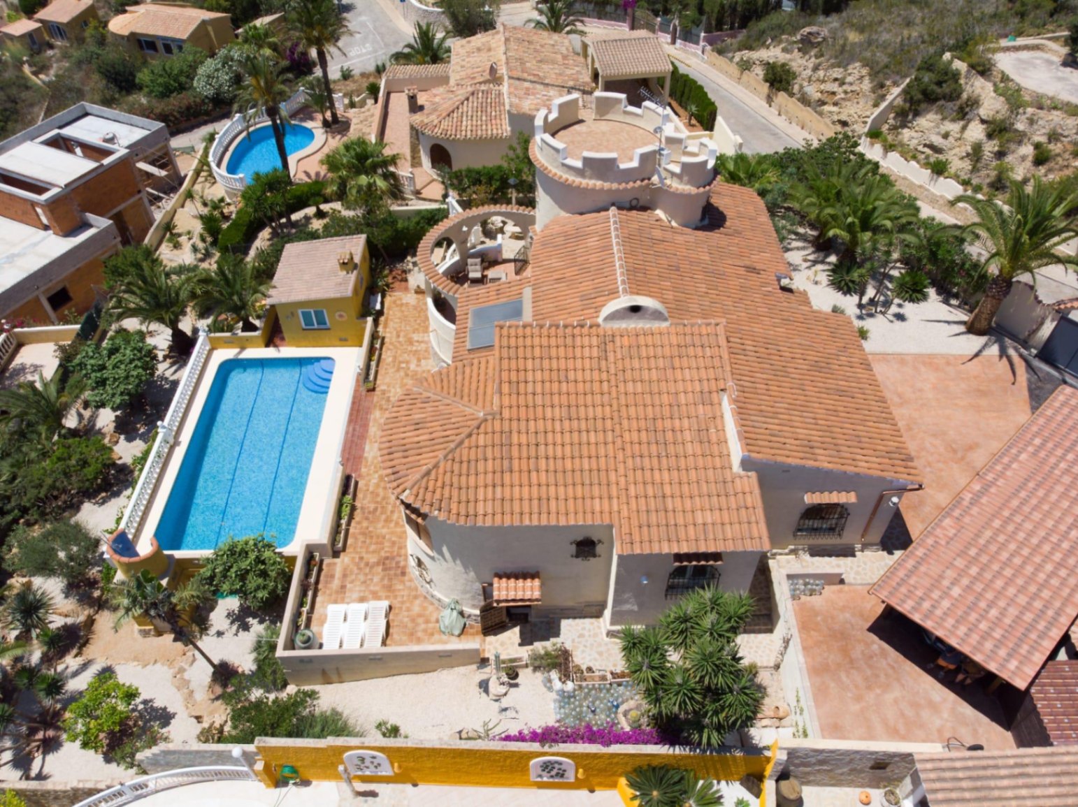 Vista aérea de la villa que muestra su arquitectura mediterránea con techos de teja, una torre distintiva y una piscina rectangular rodeada de terrazas y vegetación.