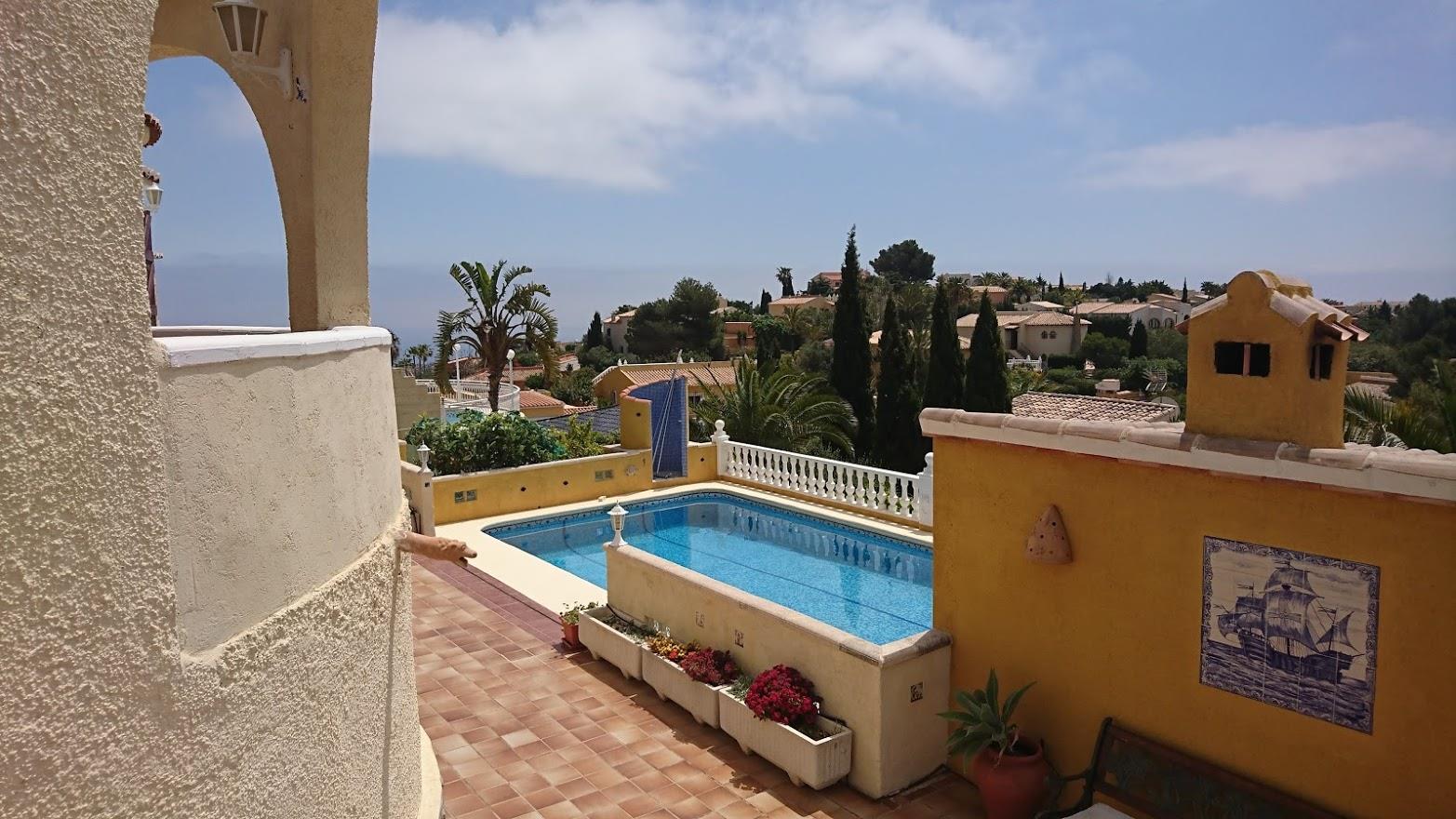 Vista elevada de la piscina rectangular rodeada de una terraza de terracota, muros ocres con detalles cerámicos y vegetación mediterránea con vistas al mar.