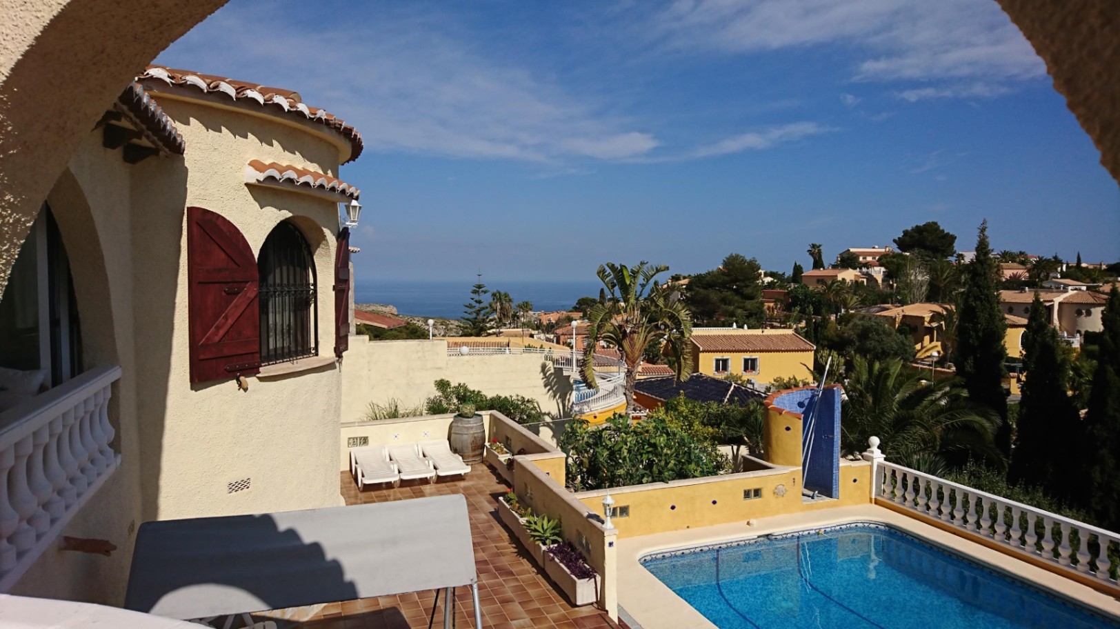 Vista exterior con piscina rectangular, terraza de terracota con tumbonas y torre arquitectónica con contraventanas de madera y vistas al mar.
