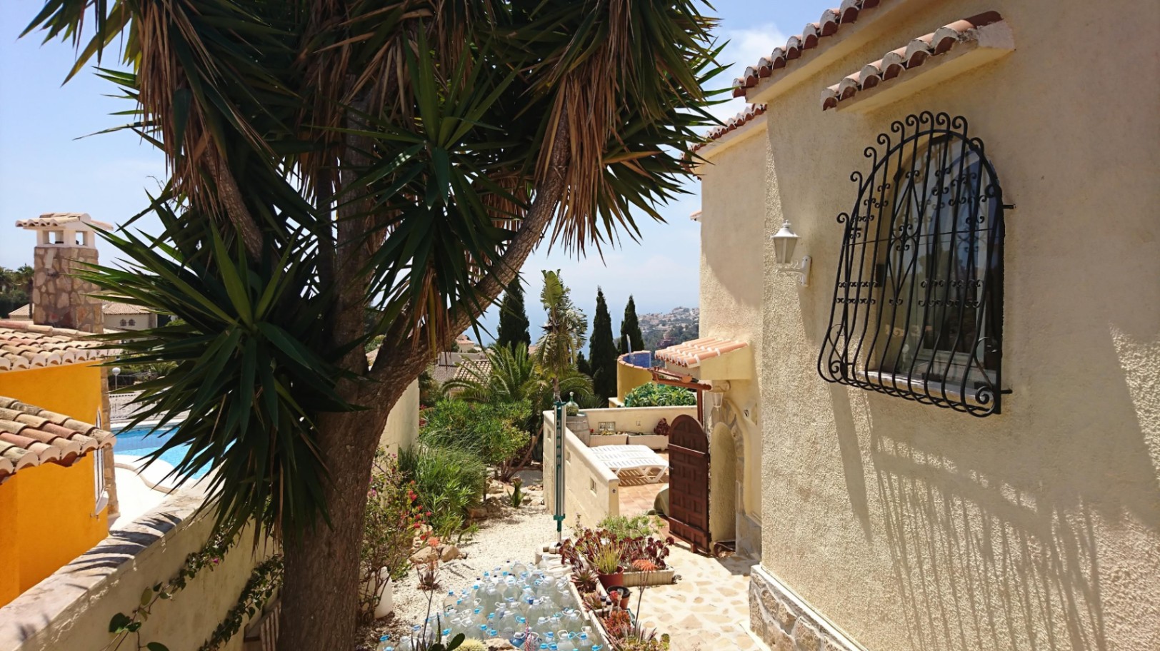 Vista exterior de la villa con fachada de mortero rugoso, ventana protegida por reja de hierro forjado y jardín mediterráneo con vistas al mar.