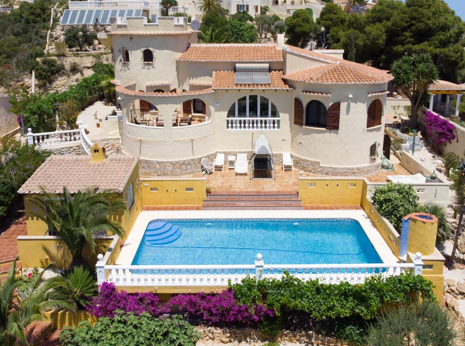 Vista exterior de la villa con piscina rectangular, terraza de terracota y fachada con torre característica, rodeada de vegetación mediterránea y muros amarillos.
