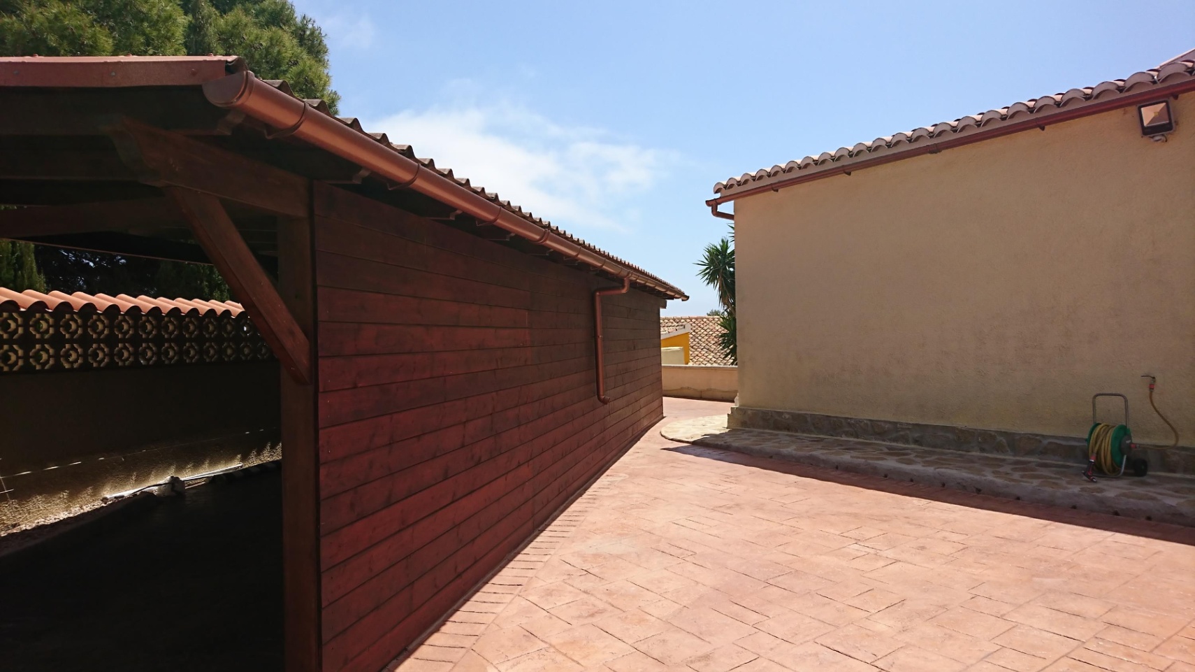 Zona exterior pavimentada con hormigón impreso que conecta un garaje de madera con la fachada principal, bajo un cielo despejado.