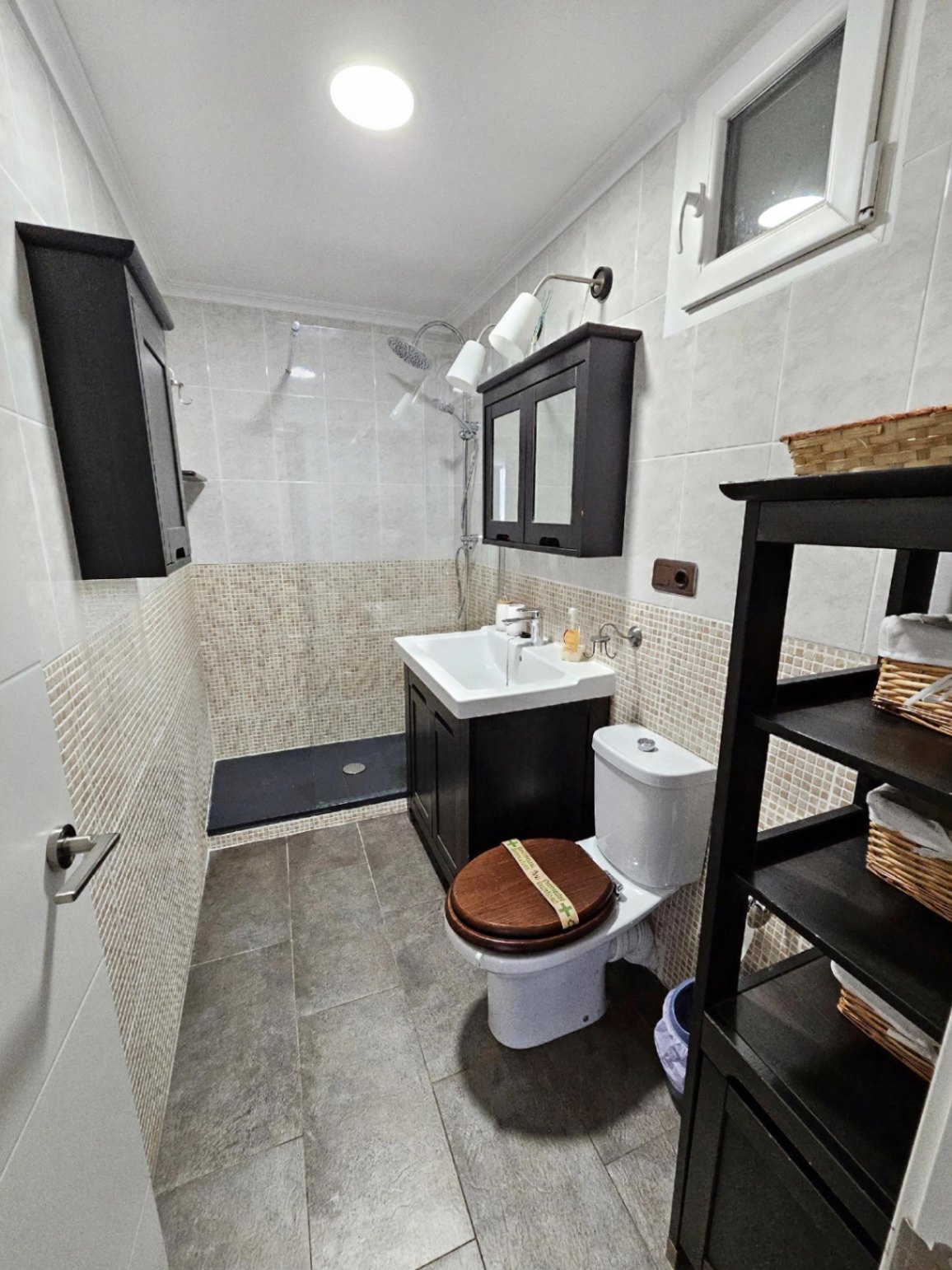Baño reformado con ducha a ras de suelo, paredes revestidas de mosaico en tonos neutros y mobiliario de madera oscura con lavabo integrado.