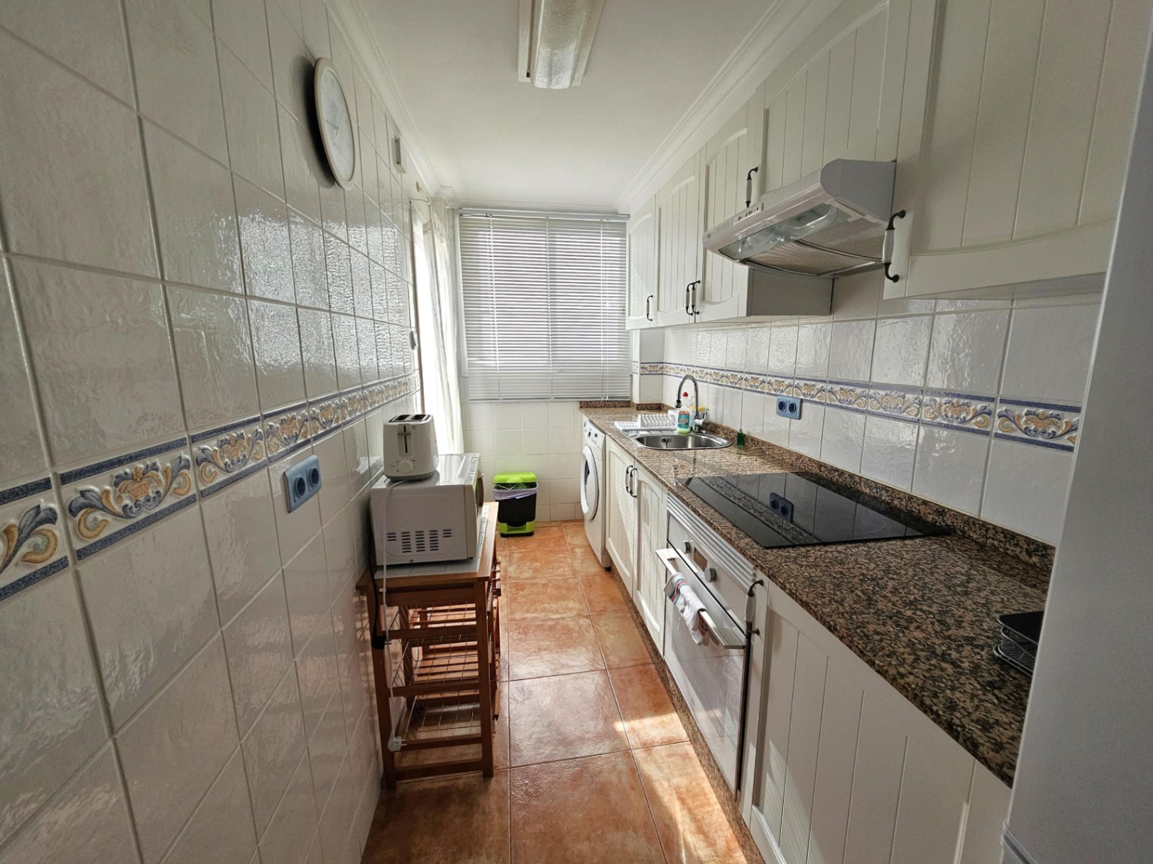 Cocina independiente de diseño alargado con encimera de granito, armarios blancos y equipamiento completo que incluye vitrocerámica, horno y zona de lavadero al fondo.