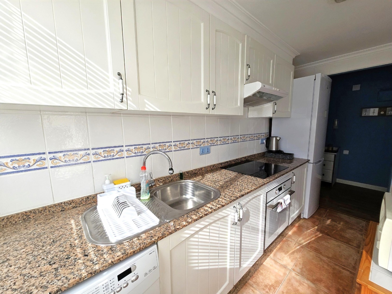 Cocina reformada con armarios blancos de estilo rústico, encimera de granito y electrodomésticos integrados. Incluye un frente de azulejos con cenefa decorativa y suelo cerámico.