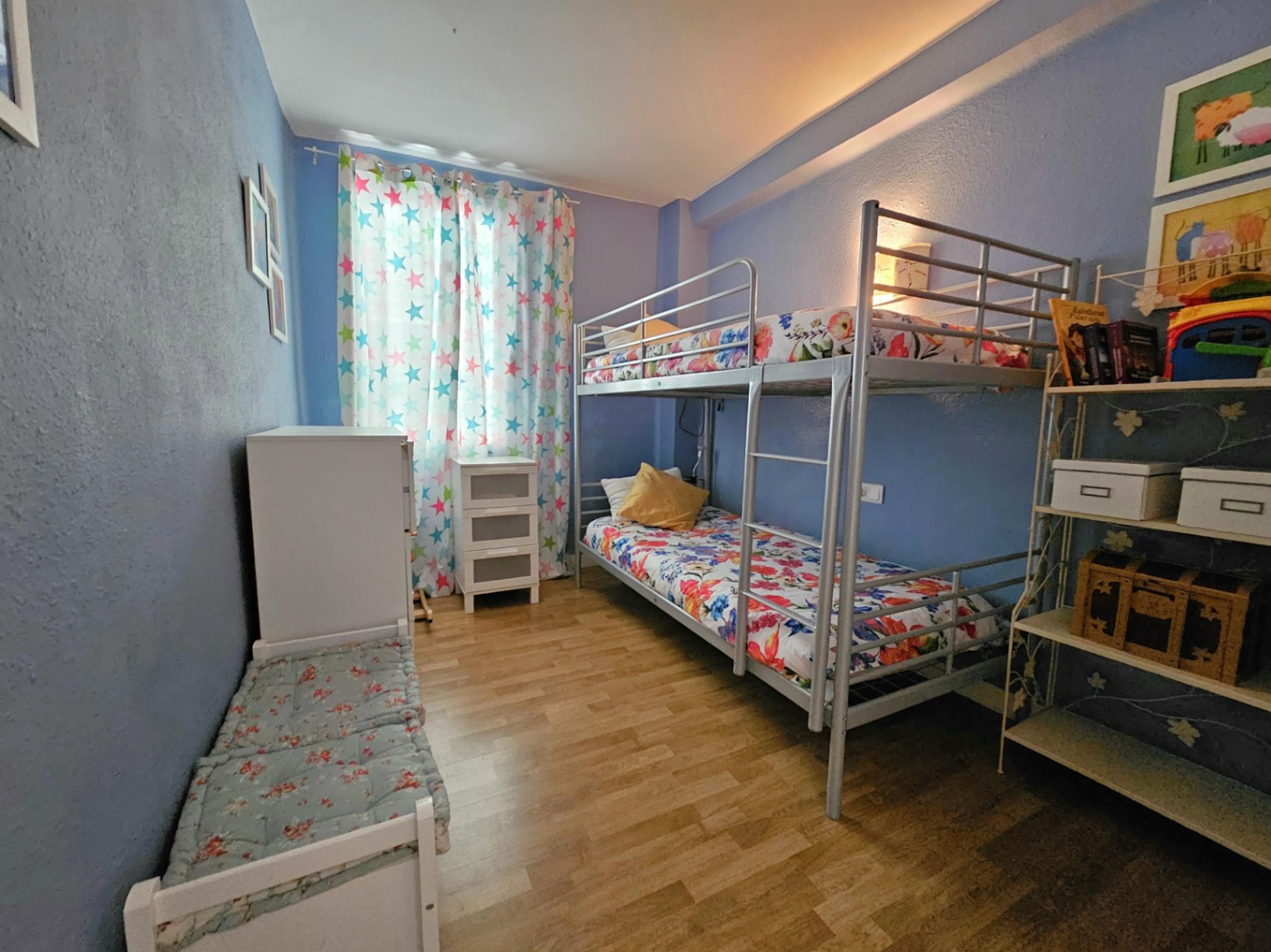 Dormitorio con litera metálica, suelos de parqué y paredes en tono azul. Incluye mobiliario auxiliar blanco y estanterías, con luz natural a través de cortinas estampadas.