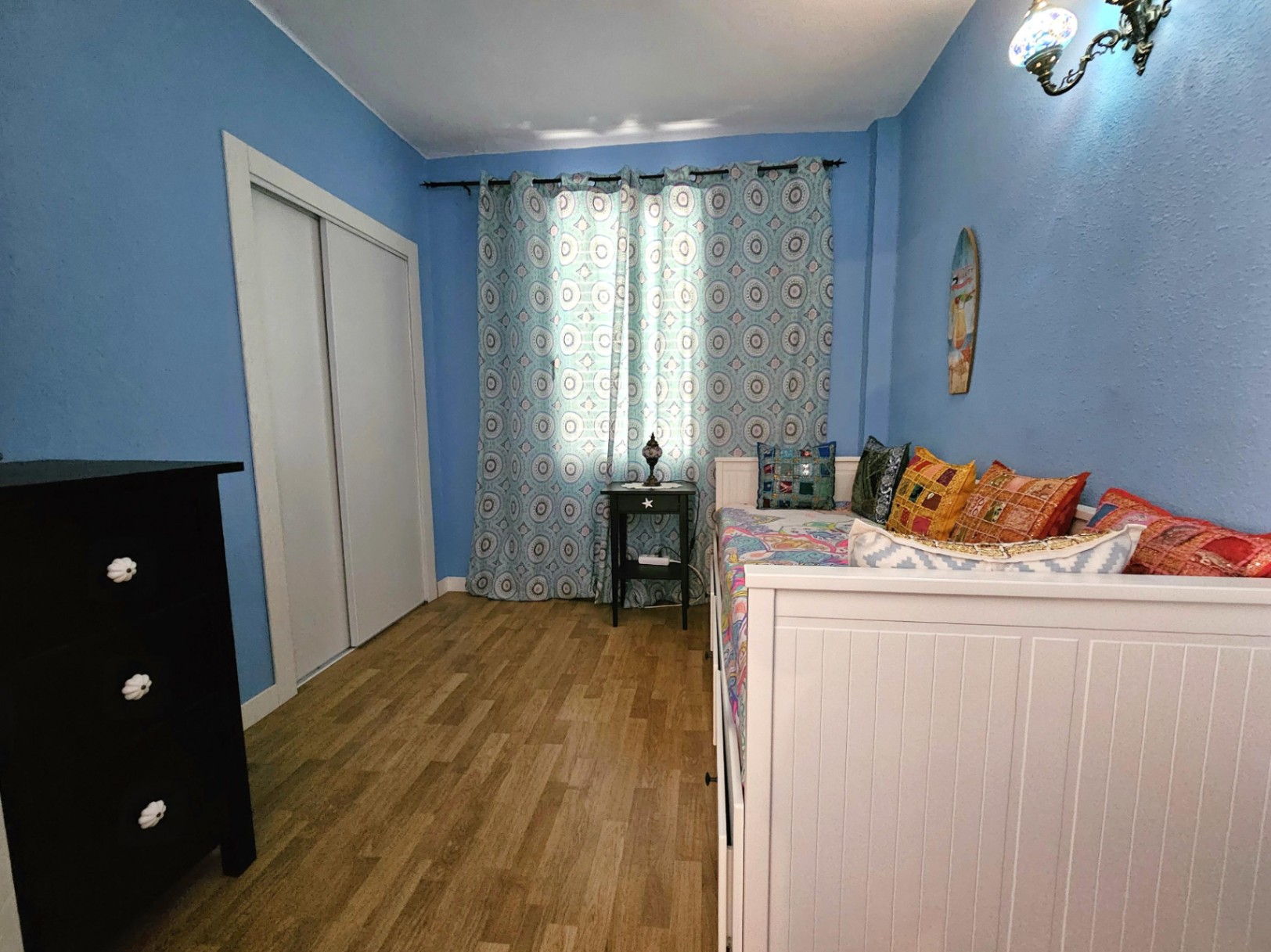 Dormitorio con paredes azules, suelo de parqué, armario empotrado blanco y una cama nido con cojines coloridos junto a una cómoda negra.