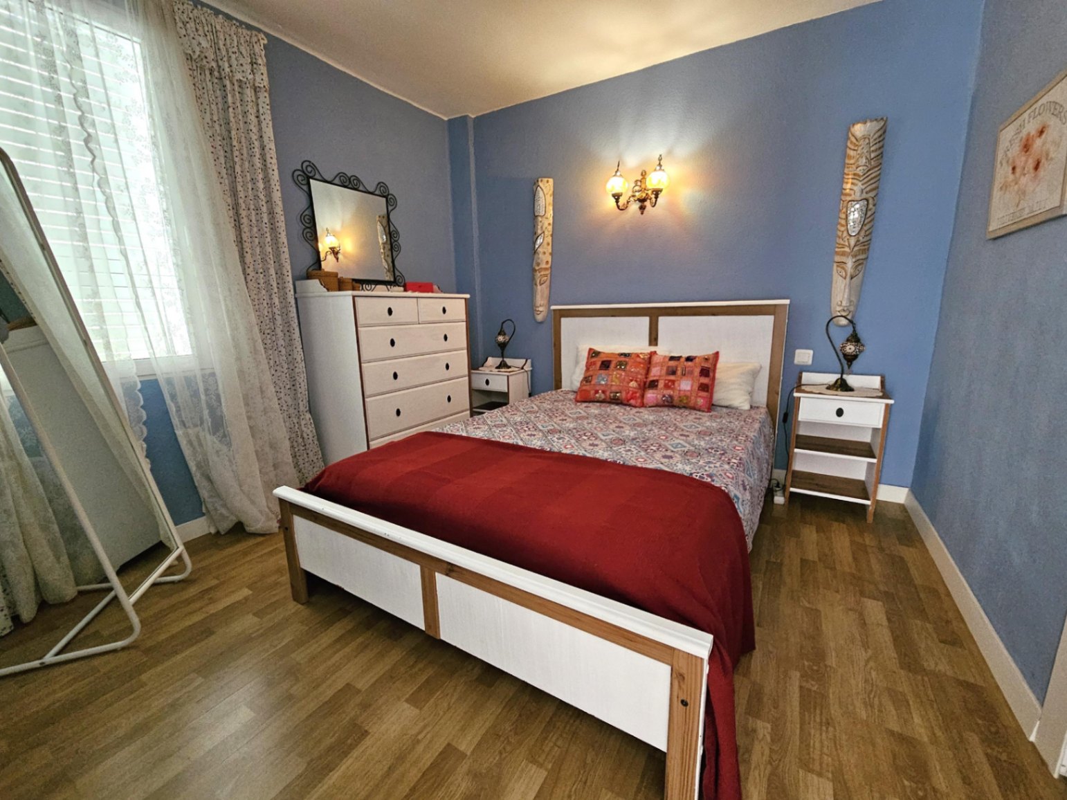 Dormitorio con paredes en azul suave, suelo de parqué y mobiliario de madera blanca. Dispone de cómoda, espejo de pie y luz natural filtrada por cortinas de encaje.