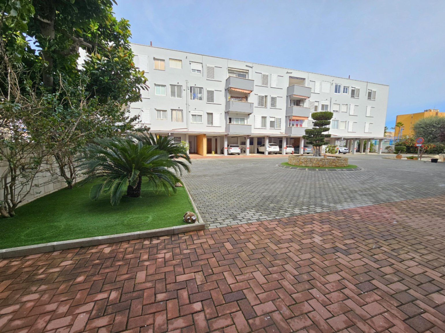 Exterior del complejo residencial con patio pavimentado, zonas ajardinadas con palmeras y plazas de aparcamiento cubiertas en la planta baja.