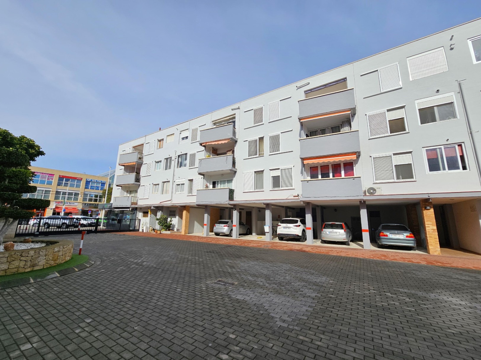 Fachada exterior del edificio con balcones y zona de aparcamiento cubierto privado en la planta baja, situada en un recinto pavimentado.