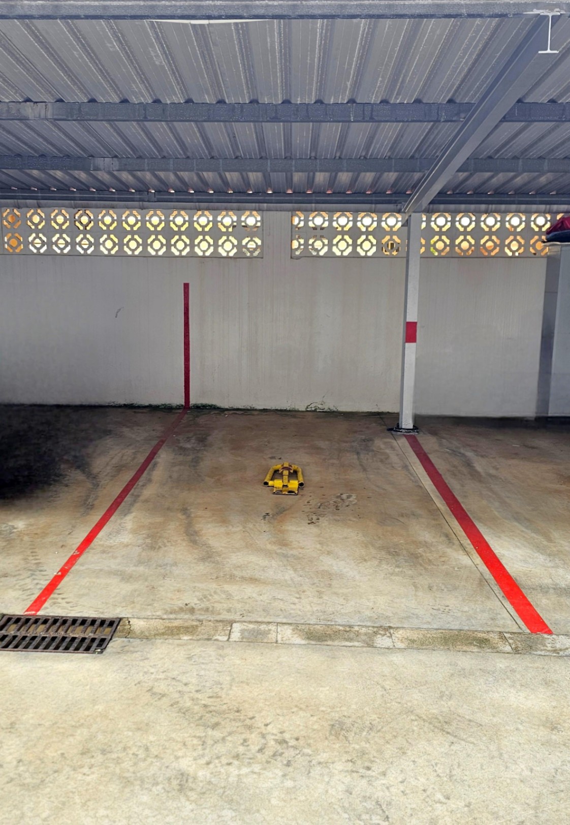 Plaza de aparcamiento privada y cubierta, delimitada por líneas rojas y equipada con un cepo de seguridad amarillo bajo una estructura de techo metálico.