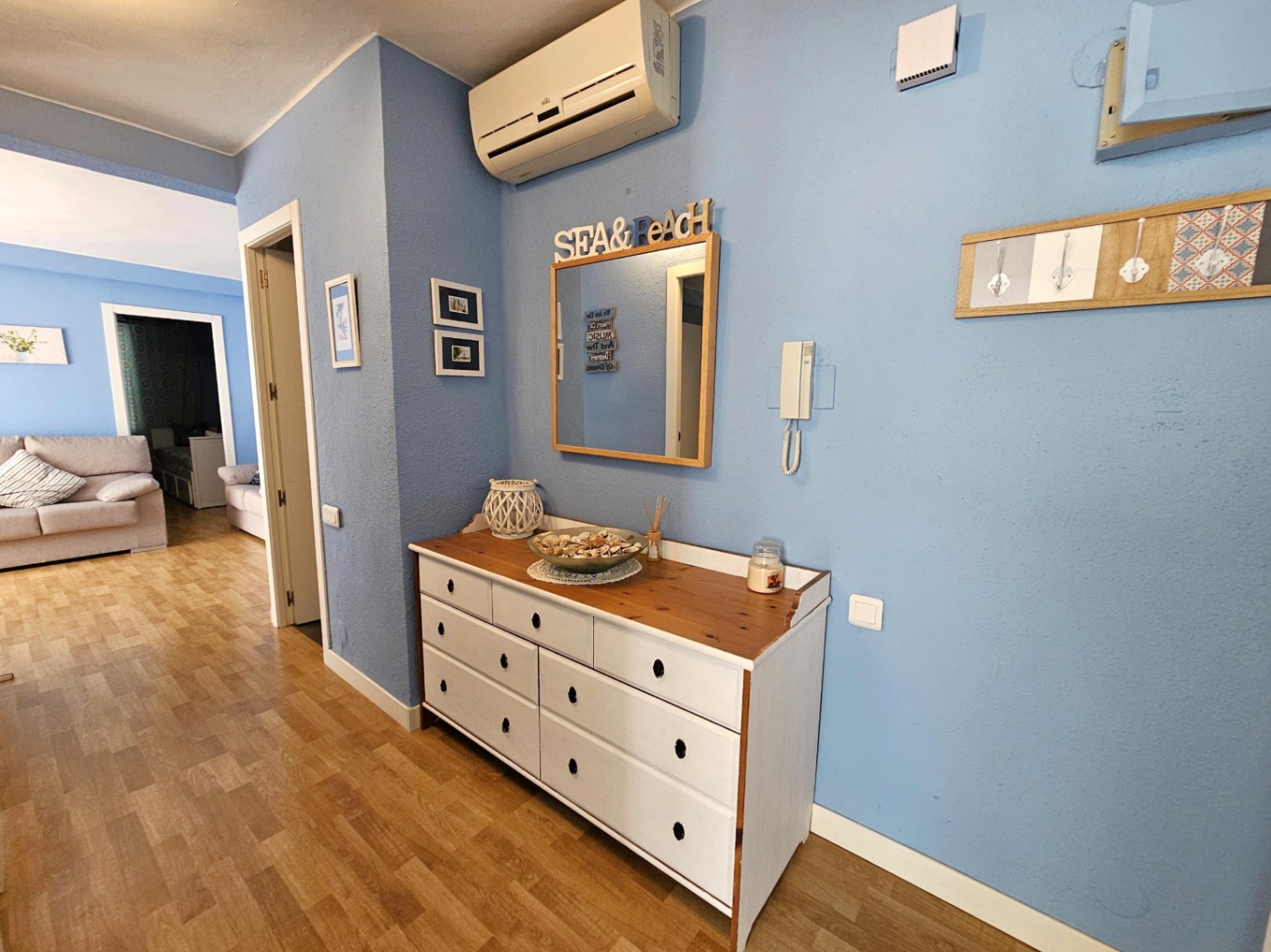 Recibidor con paredes azules y suelos de parqué, equipado con una cómoda de madera blanca, aire acondicionado y acceso directo al salón y dormitorios.