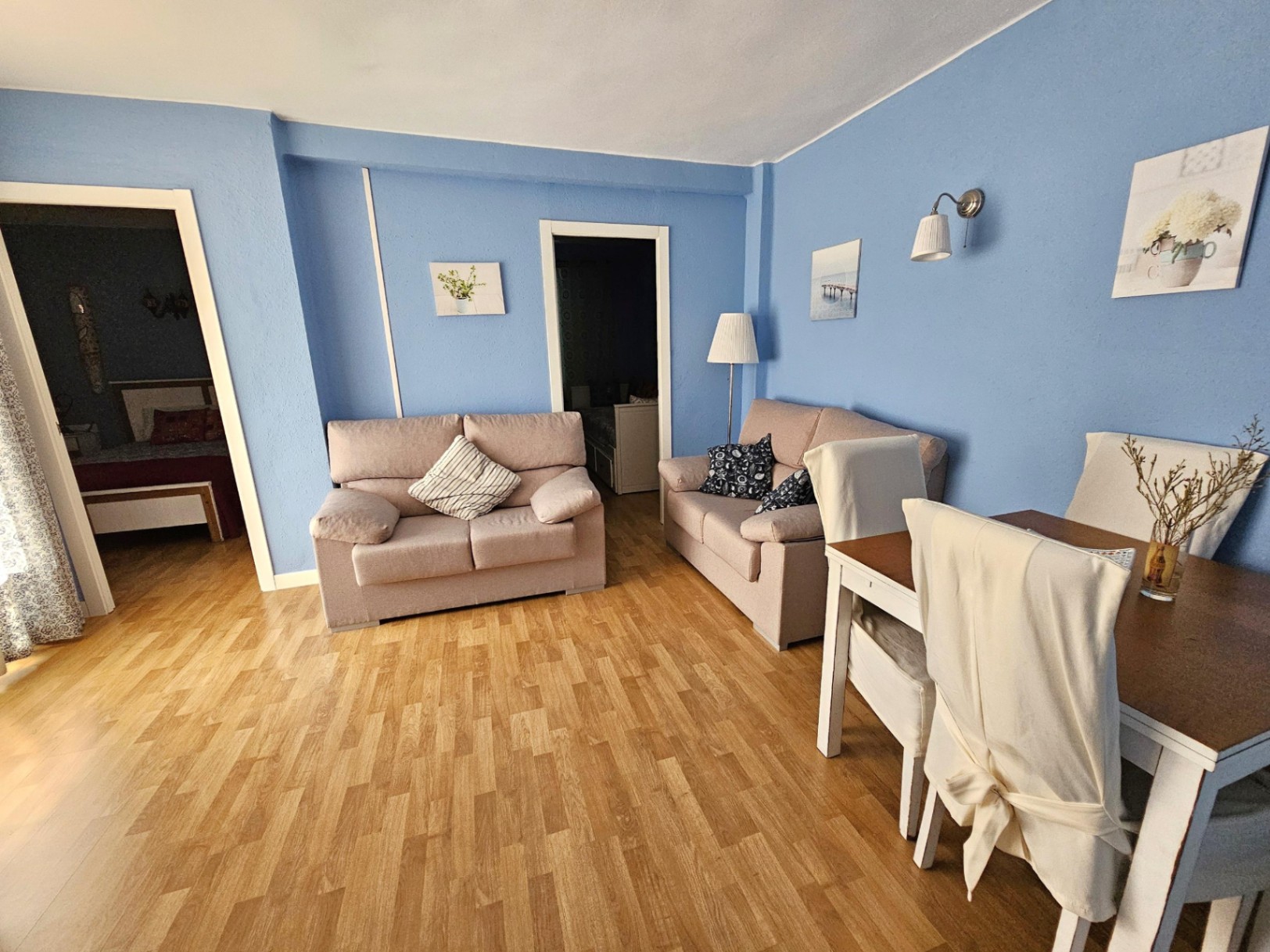 Salón comedor con paredes azules, suelos de parqué y mobiliario contemporáneo. Incluye dos sofás en tonos neutros y zona de comedor junto al acceso a los dormitorios.