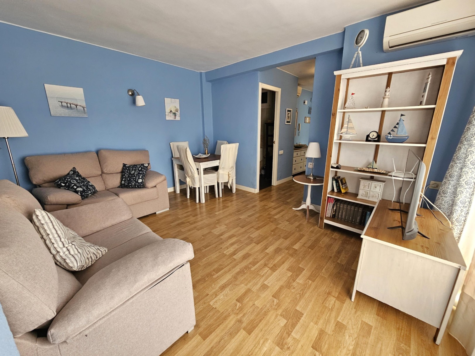 Salón comedor con paredes azules, suelos de parqué y mobiliario de estilo costero. Incluye sofá beige, zona de comedor y estantería de madera blanca.