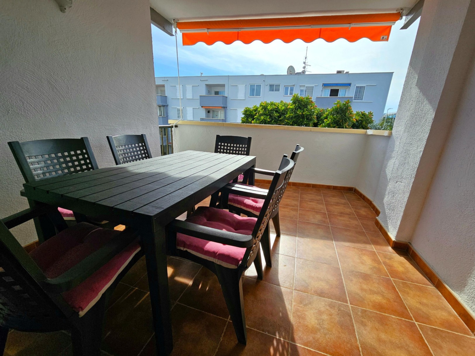 Terraza exterior con suelo de gres cerámico, equipada con un conjunto de comedor para seis comensales y un toldo retráctil de color naranja para protección solar.