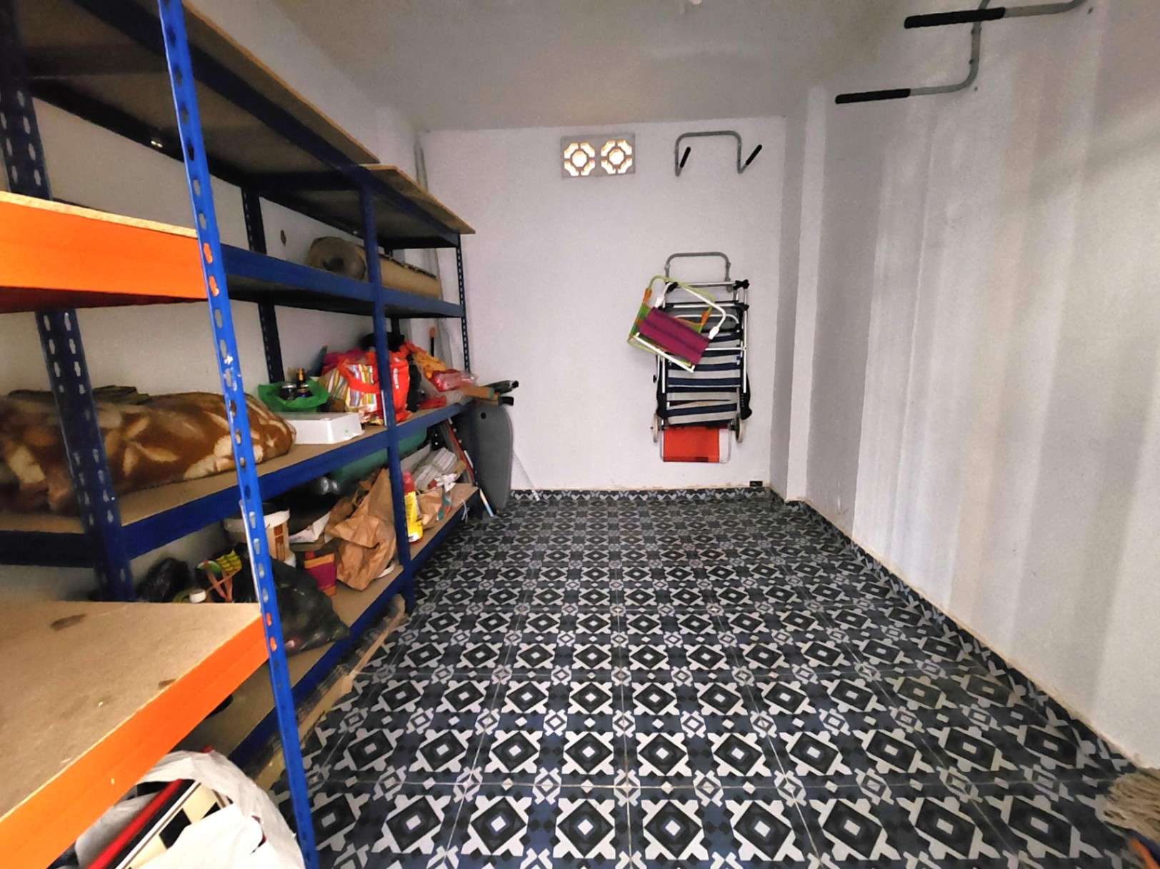 Trastero privado de 12 m² con estanterías metálicas de carga, suelo de gres con motivos geométricos y ganchos de pared para equipamiento.