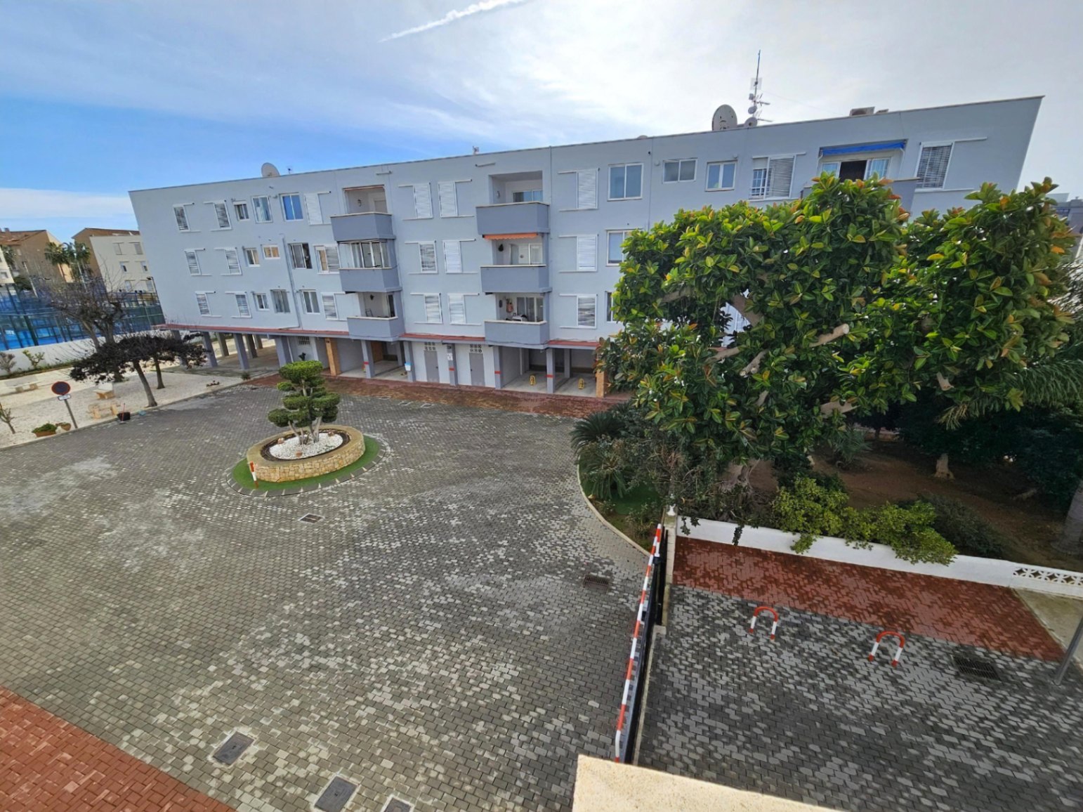 Vista exterior del complejo residencial que muestra el patio pavimentado, zonas ajardinadas y la fachada del edificio con contraventanas blancas y balcones.