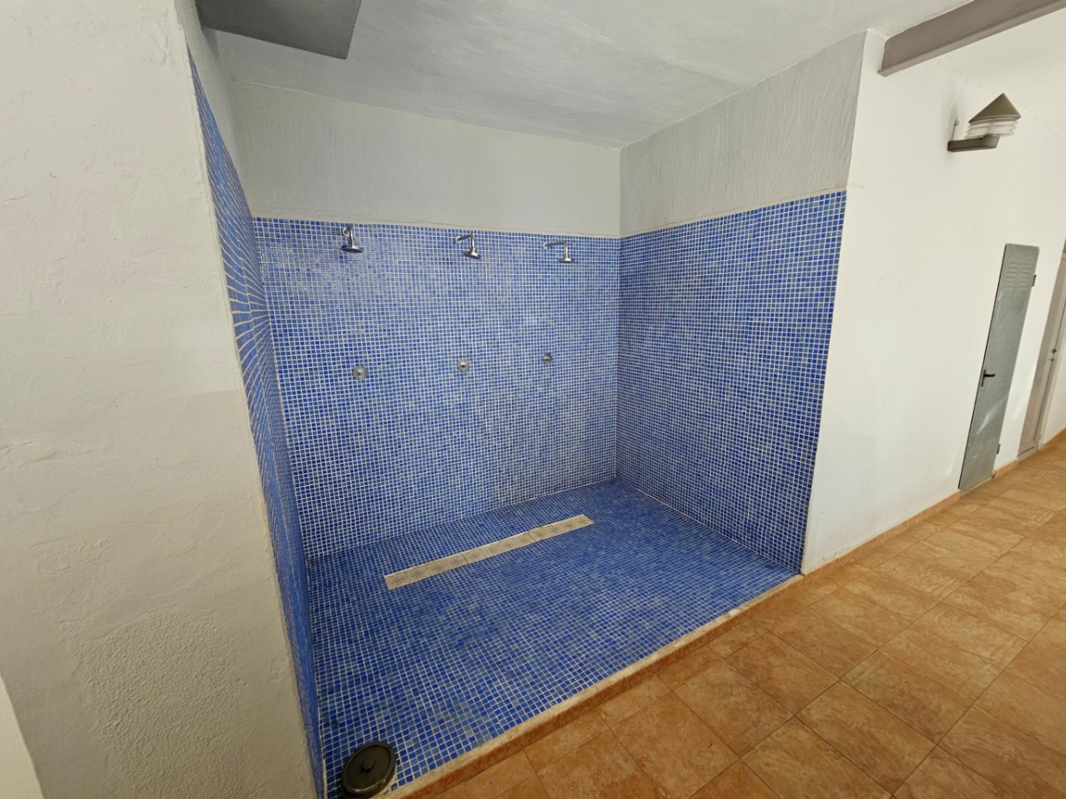 Zona de duchas comunitarias revestida con gresite azul, equipada con tres tomas de agua y desagüe central, situada en las áreas comunes del edificio.
