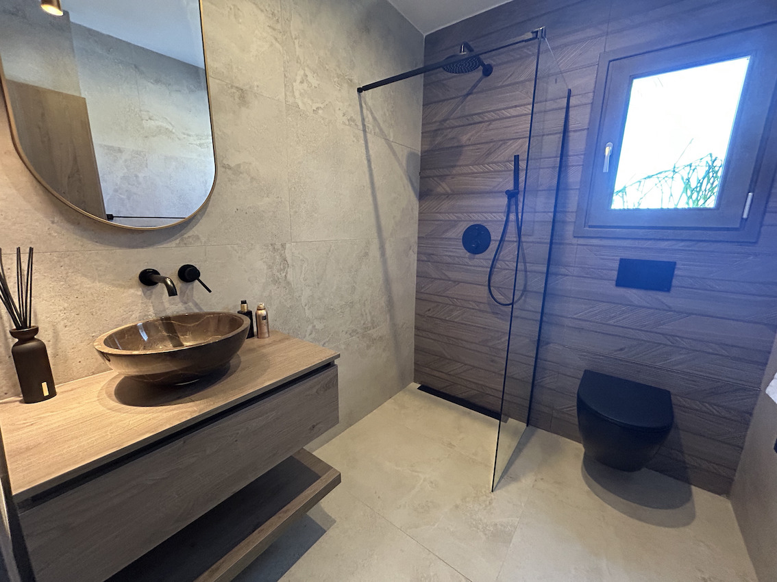 Baño moderno con lavabo de piedra sobre mueble de madera, ducha de efecto lluvia con mampara de cristal y sanitarios suspendidos en color negro mate.