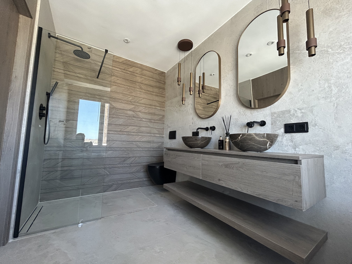 Baño moderno con lavabo doble de piedra, grifería negra empotrada, espejos ovales y ducha italiana con revestimiento cerámico de efecto madera.