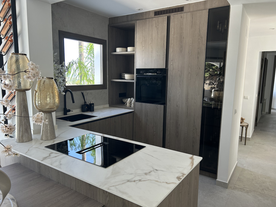 Cocina moderna con encimeras de efecto mármol, placa de inducción integrada y armarios de madera oscura. Dispone de fregadero negro mate y electrodomésticos encastrados.