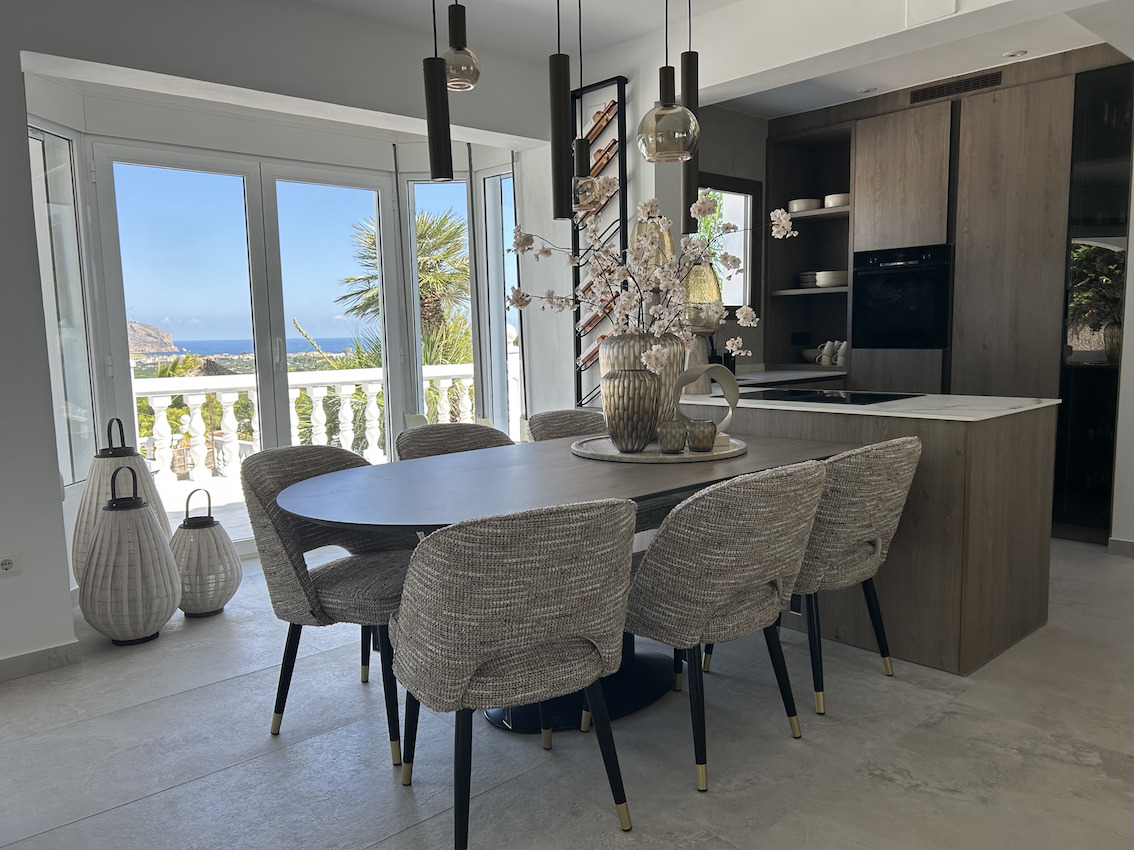 Comedor de concepto abierto con mesa ovalada y cocina integrada de diseño en madera, ofreciendo vistas panorámicas al mar a través de amplios ventanales.