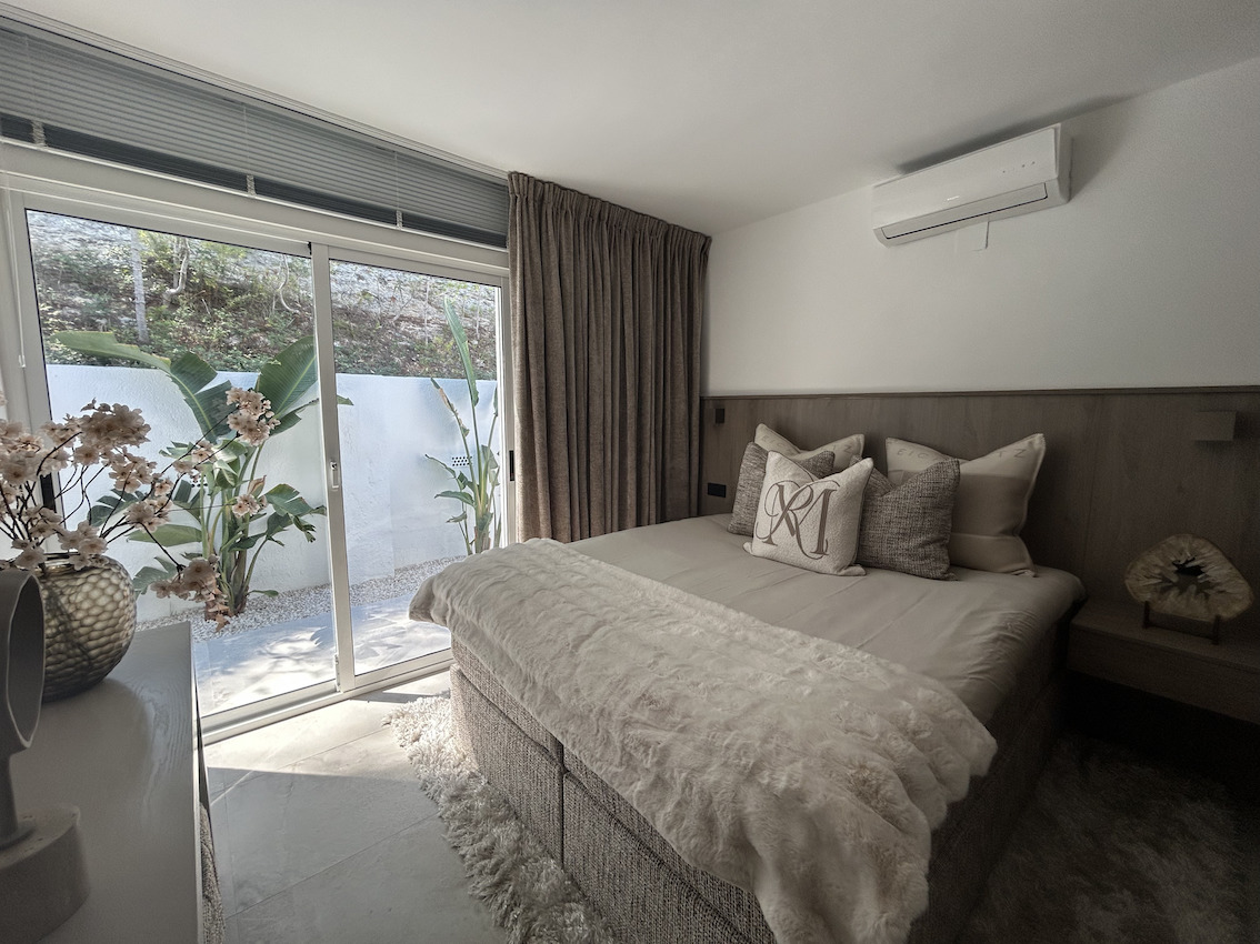 Dormitorio moderno con cabecero de madera, aire acondicionado y acceso directo a un patio privado con plantas tropicales a través de puertas correderas.