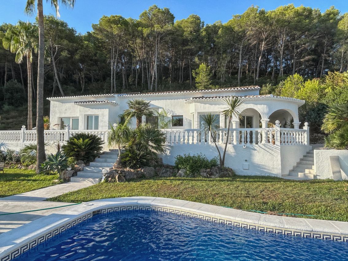 Fachada blanca de la villa con terraza de balaustrada, jardín mediterráneo y piscina en primer plano, rodeada de un frondoso bosque de pinos.
