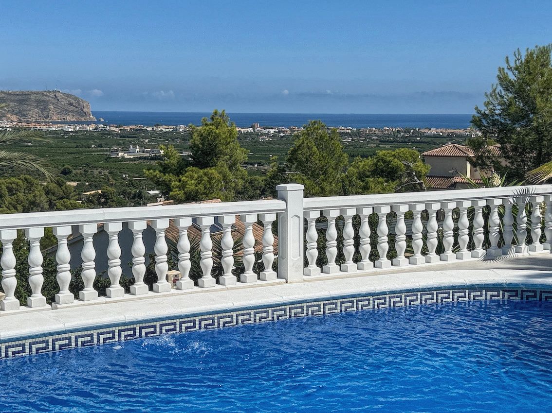Piscina con borde de mosaico azul y balaustrada blanca, ofreciendo vistas panorámicas al mar Mediterráneo, el valle y la costa bajo un cielo despejado.