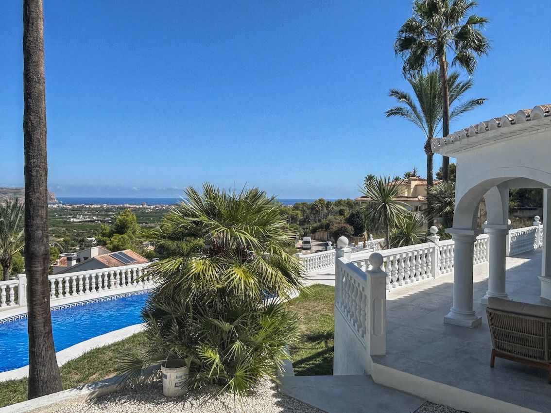 Terraza con balaustrada blanca y porche de arcos que ofrece vistas panorámicas al mar y al valle, rodeada de palmeras y zona de piscina.