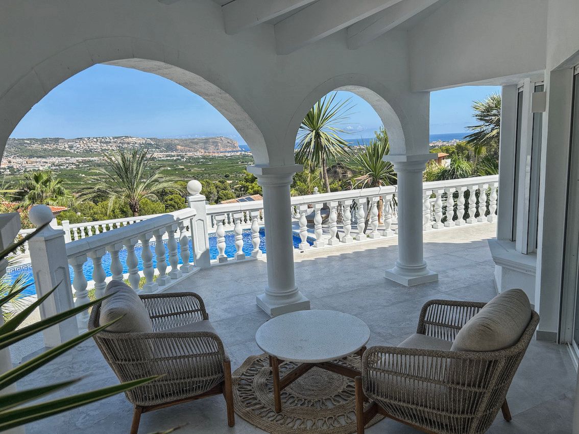 Terraza cubierta con arcos blancos y columnas, amueblada con sillones de mimbre. Ofrece vistas panorámicas al valle, el mar y la piscina privada.