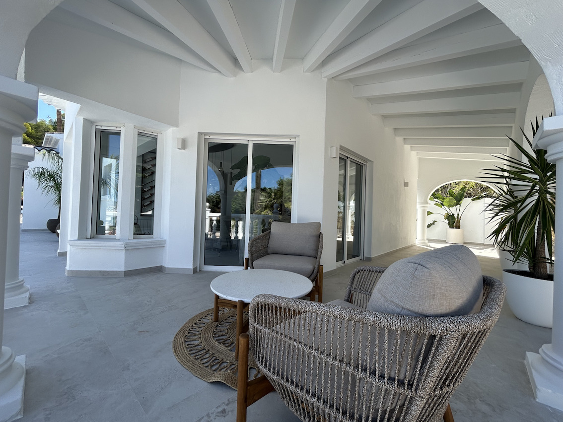 Terraza cubierta con vigas de madera blancas, mobiliario de exterior moderno y suelos de gres porcelánico, conectada al interior mediante puertas correderas.